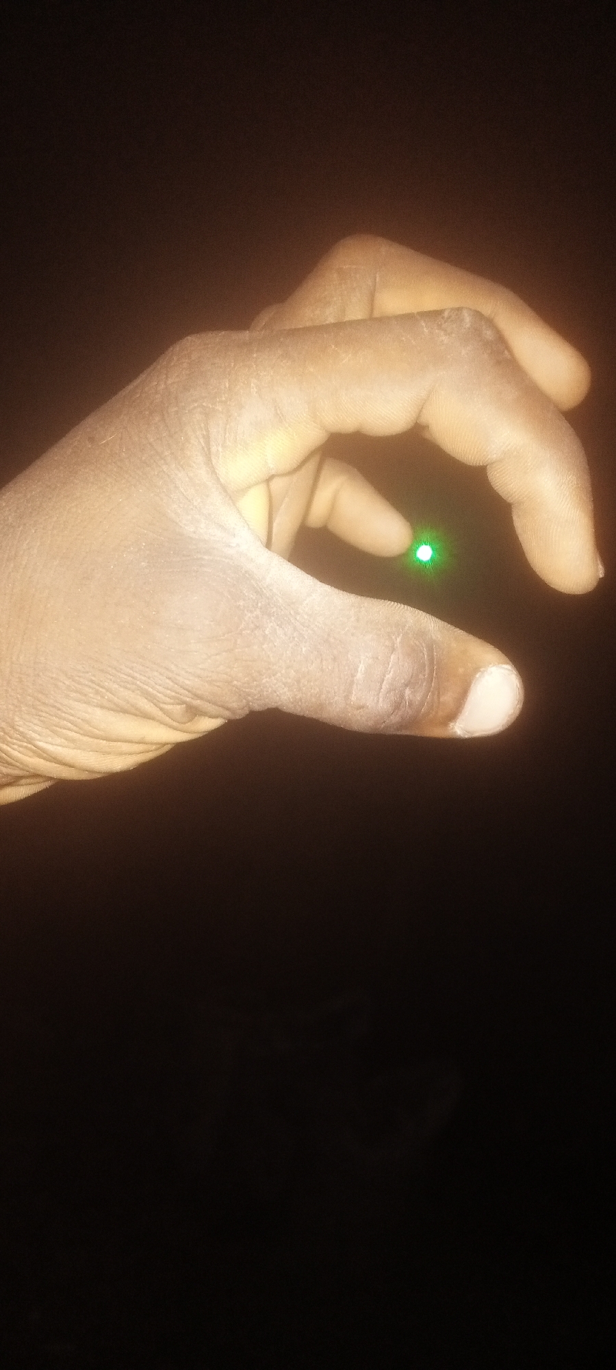 A right hand with dark skin is holding a green laser pointer between the index finger and thumb. The image is set against a dark, blurry background. The location in the city of Narayi, Nigeria is undetermined.