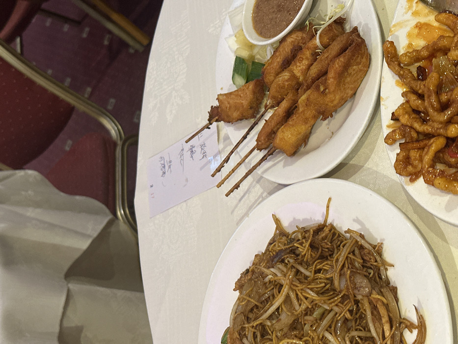 Three plates of food sit on a white tablecloth. One plate holds fried noodles with visible sprouts and onions. Another plate contains meat skewers with a dipping sauce. The third plate is filled with a sticky, orange-glazed meat. A piece of paper with handwritten text lies partially under the plate of skewers. A blurred chair with a red cushion is visible in the background.