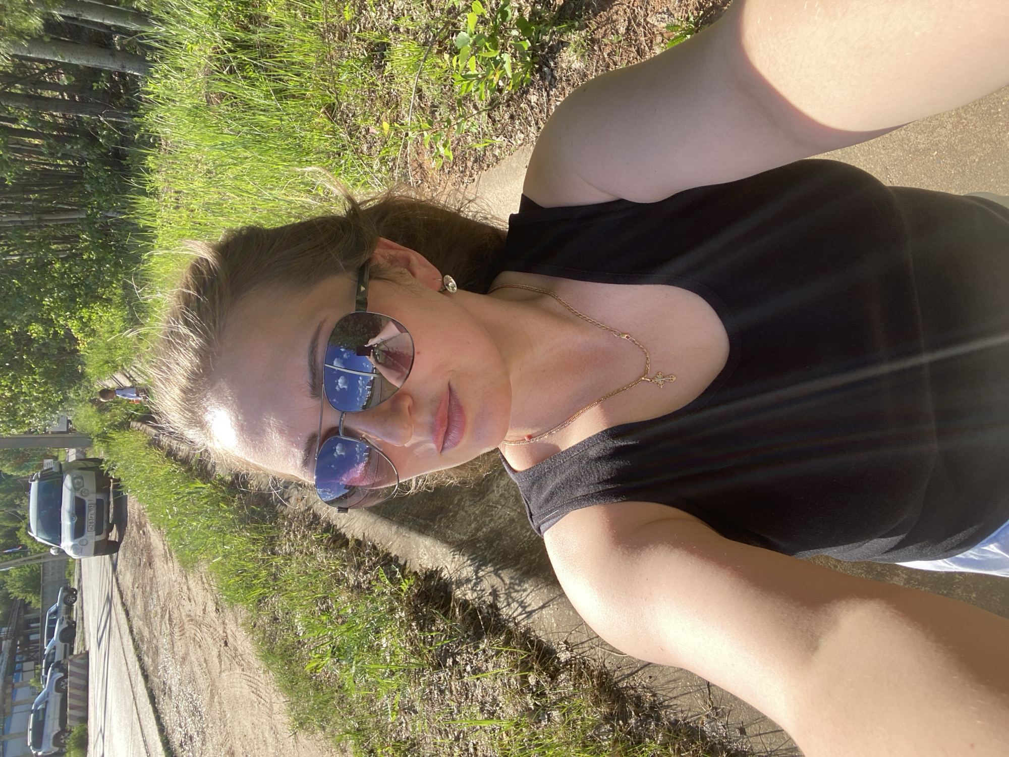 A selfie taken in Chaikovsky, Russia, features a fair-skinned woman wearing sunglasses, a black tank top, and a gold necklace with a cross pendant. She is positioned on a paved path with grass growing along the edge. In the background, there's a dirt road with parked cars and a white SUV bearing the license plate "H213PH 161". The landscape includes trees and glimpses of buildings. The sunglasses reflect the sky, indicating daylight conditions. The angle of the photo suggests it was taken with a handheld device.