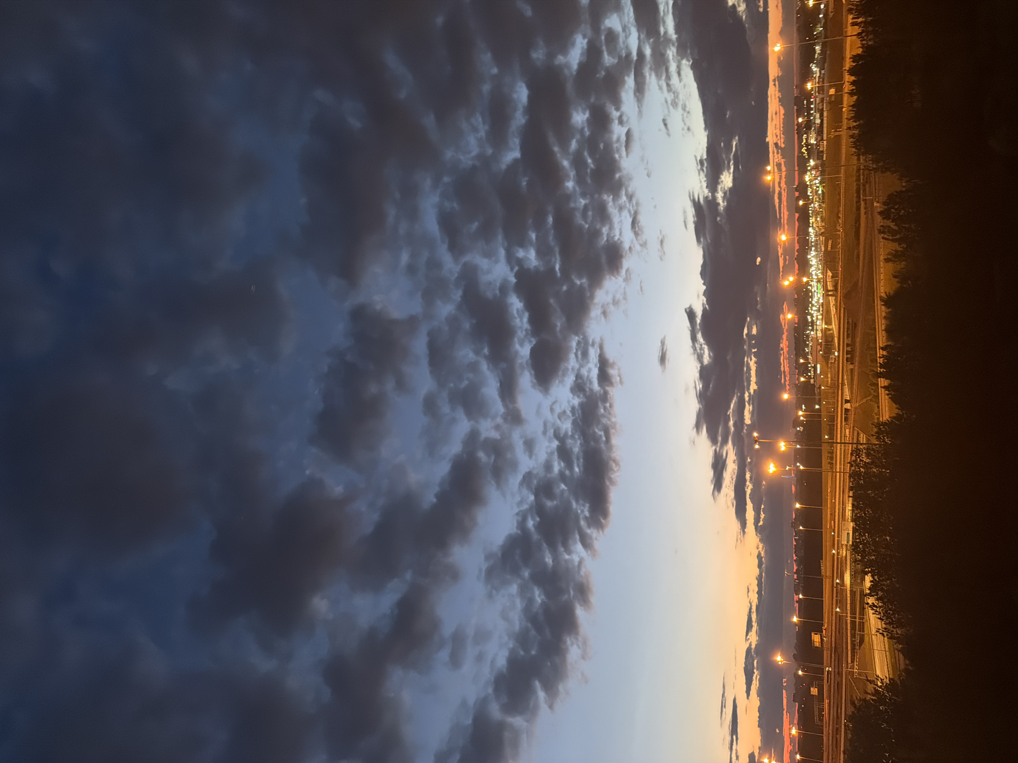 The image depicts an urban landscape at dusk or dawn. The sky is a gradient of colors, transitioning from a deep blue with dark clouds at the top to lighter blues and oranges near the horizon. A dense arrangement of clouds fills the upper portion of the frame. In the lower half, a sprawling network of roads is illuminated by numerous streetlights, casting a warm glow. Beyond the roads, a cityscape with buildings is visible, further lit by artificial lights. Silhouetted trees form a dark band across the bottom of the image, obscuring the immediate foreground. The overall impression is one of a city awakening or settling down under a dramatic sky.