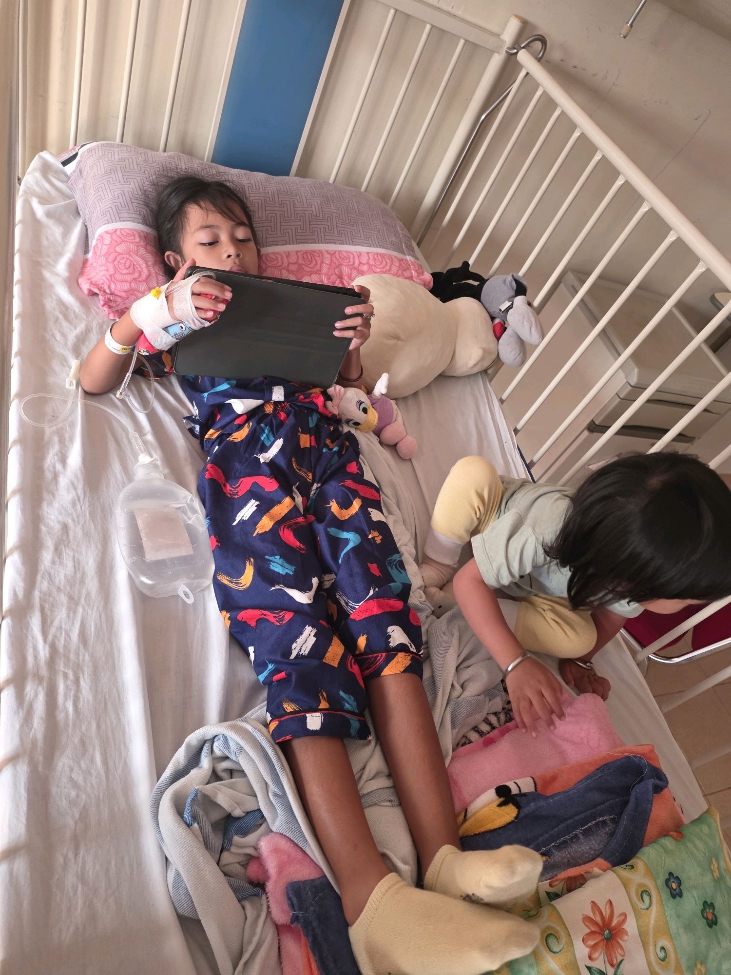 Two young children are lying on a hospital bed. The child on the left is propped up on pillows and is looking at a tablet device. They have an IV drip connected to their arm. The child on the right is sitting up and appears to be reaching for something on the bed. The bed has a metal railing on one side. There are several stuffed animals on the bed, as well as blankets and pillows. The room appears to be a hospital room, with a white bedsheet and beige walls. The children are wearing casual clothing. This image was taken in a hospital in Tanjung Pinang, Indonesia.