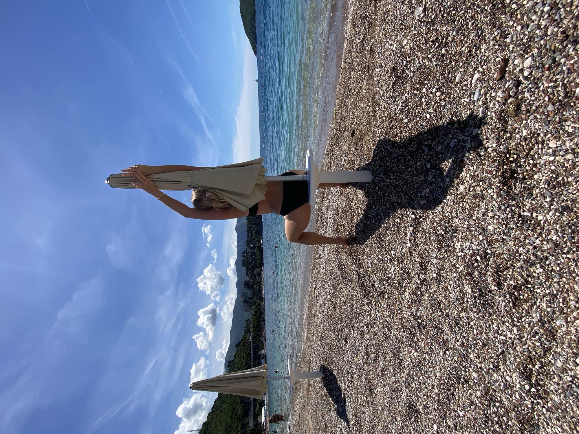 The image depicts a sun-drenched beach scene, likely in Budva, Montenegro, under a clear blue sky with scattered white clouds and faint contrails. The shore is composed of small, multicolored pebbles and gravel, meeting tranquil, blue-green water.
In the foreground, a person, viewed from behind, is the primary subject. Dressed in a black two-piece swimsuit, they are bending forward, holding the top of a closed, light-colored (beige or tan) beach umbrella with both hands, appearing to be in the process of adjusting or opening it. The umbrella's white pole is visible, planted in the pebbles. The person casts a strong, distinct shadow on the pebbled ground, indicating bright sunlight.
Further down the beach, another similar closed beach umbrella stands near the water's edge. Across the calm bay, lush green mountains rise, featuring scattered buildings along their slopes. Several small objects, possibly buoys or distant boats, are visible in the water. The scene conveys a bright, sunny day at a picturesque Mediterranean beach. No visible text is present in the image.