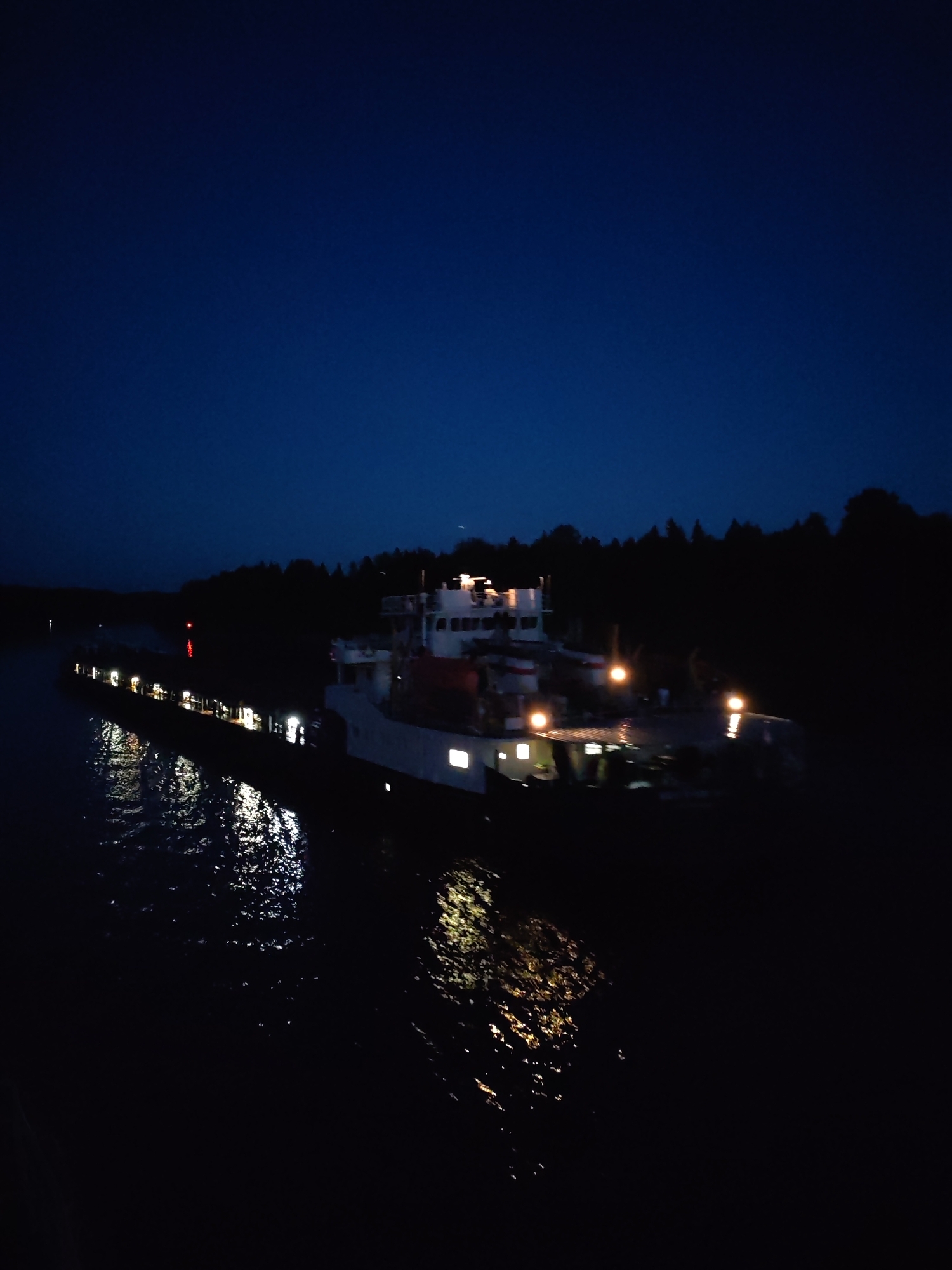 A large barge is illuminated at night on a body of water in Uzhla, Russia. The barge's structure includes a taller section towards the front, likely housing the bridge and crew quarters, and a long, lower cargo area trailing behind it. Several lights shine from the barge, reflecting in the water. Trees line the shore in the background, silhouetted against the night sky.