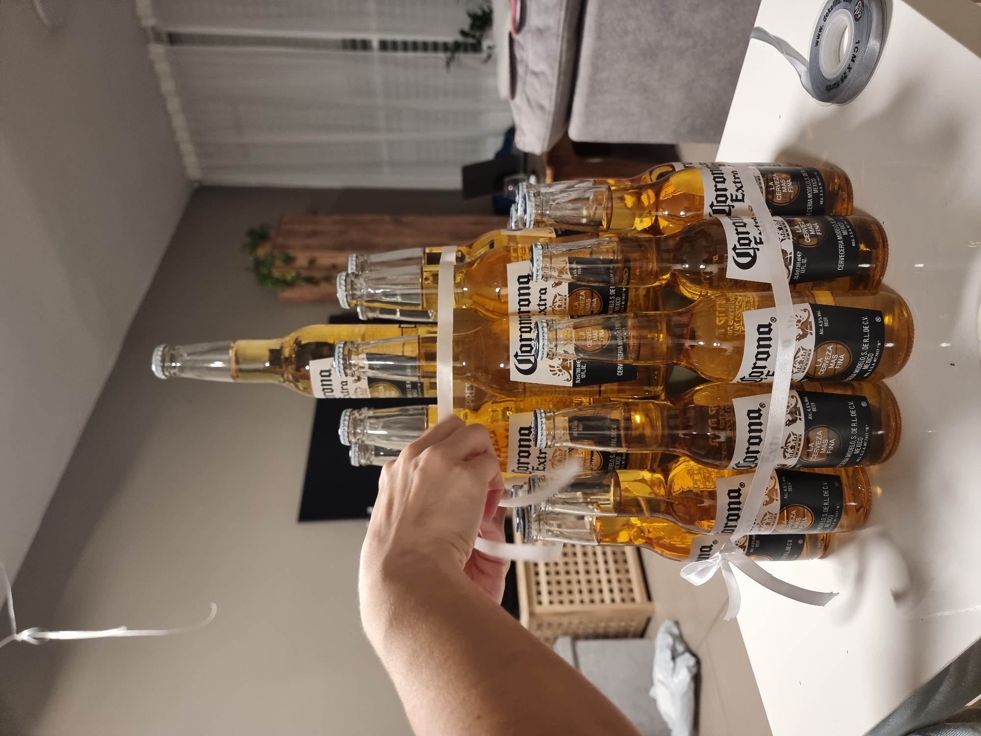 A pyramid of Corona Extra beer bottles is arranged on a white table. The bottles are tied together with a white ribbon. A person's left hand is visible, holding a length of the ribbon. In the background, a woven basket, a grey sofa, and sheer curtains can be seen. The overall lighting suggests an indoor setting, possibly a home. The image appears to have been taken in Ramat Gan, Israel.