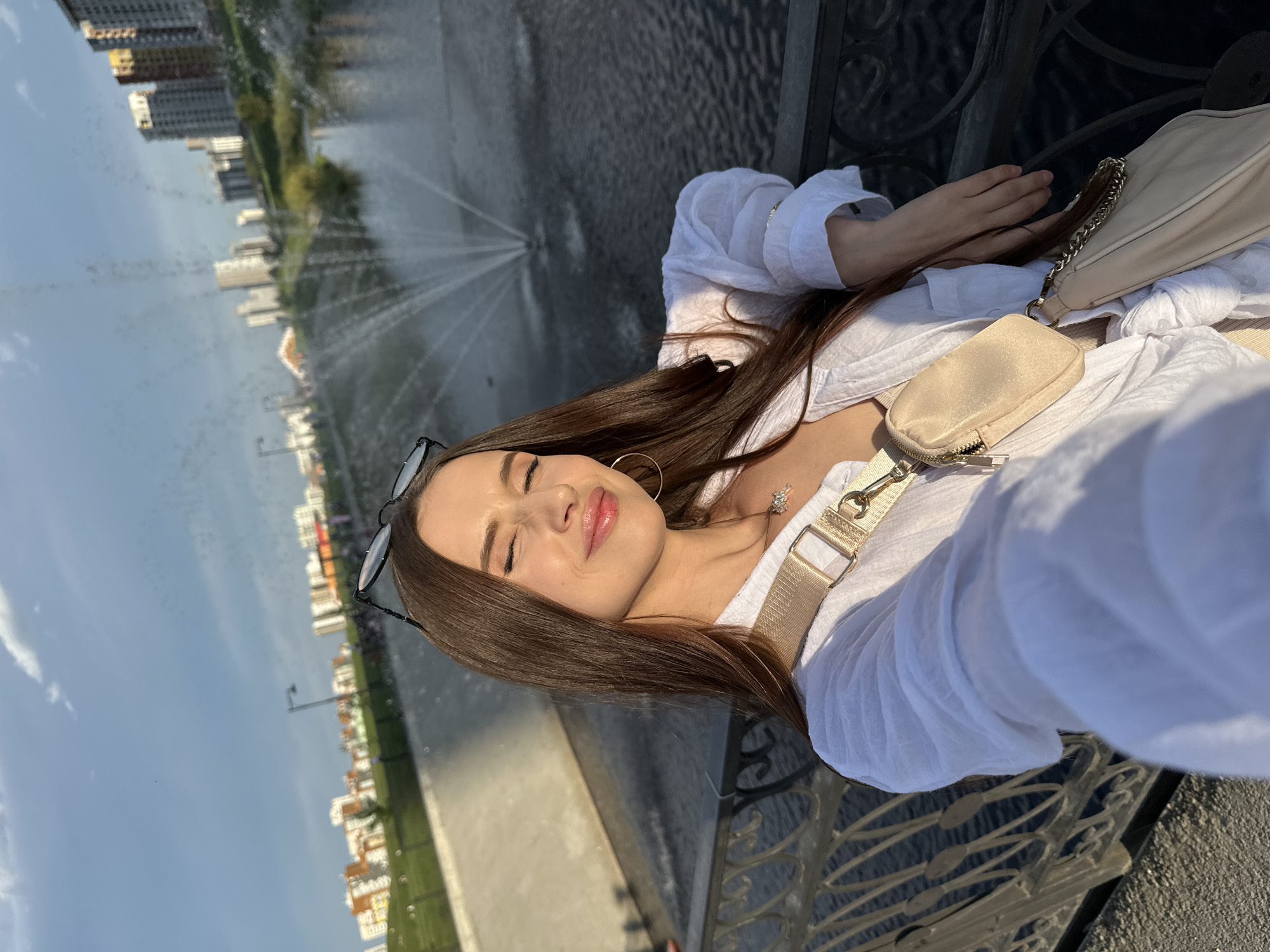 A young woman with long, brown hair and glasses is taking a selfie. She is wearing a white shirt, hoop earrings, and a crossbody bag. She is standing near a fountain, with buildings in the background. The fountain appears to be in a city, possibly in Yekaterinburg, Russia. The location is likely the "Plotinka" area, a historical dam and a popular spot in the city. The woman has her eyes closed and is smiling slightly.
