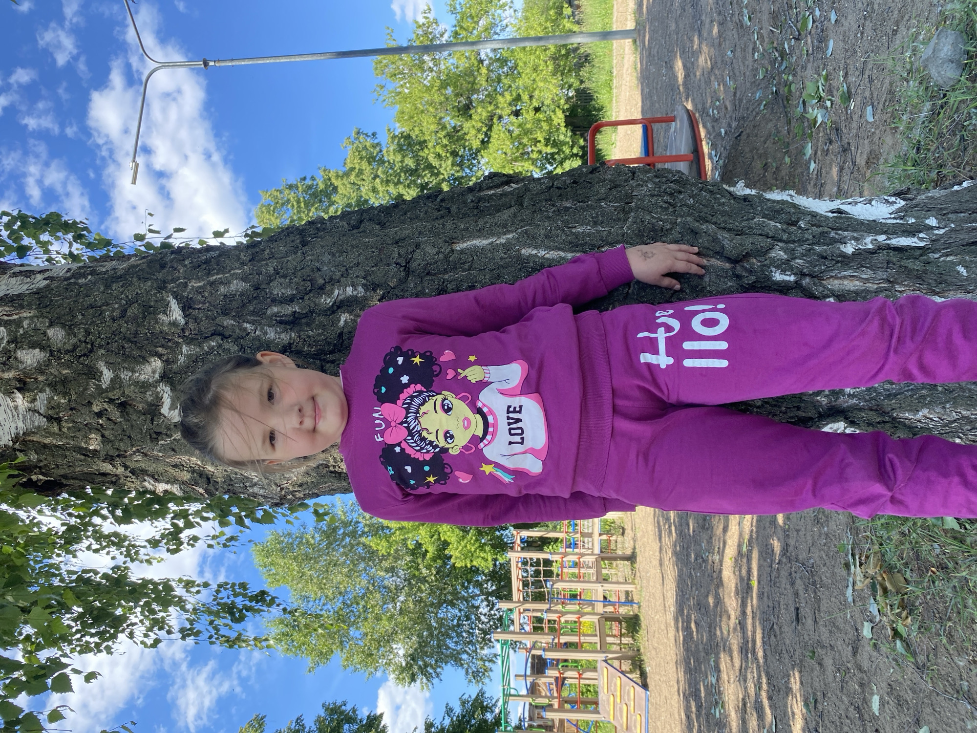 A young girl, approximately 5-8 years old, stands vertically centered in the frame, leaning her right side against a tree trunk. She is wearing a matching magenta tracksuit, consisting of a long-sleeved top and pants. The top features a graphic of a cartoon girl with dark, curly hair, a pink bow, stars, hearts, and the words "FUN" and "LOVE". The left leg of the pants has white text, possibly "Hullo!" or "Hello!". Her light brown hair is pulled back, and she is looking directly at the camera with a slight smile.
The prominent tree trunk, exhibiting bark characteristic of a birch with white patches, occupies the right side of the image from bottom to top. Green leaves hang from its branches in the upper left corner. The ground consists of dirt and sparse green grass, with fallen leaves and small rocks. Shadows from foliage are cast on the ground.
In the background on the left, a multi-colored children's playground structure is visible, featuring various climbing elements, slides, and nets. To the right of the playground and behind the girl, more green trees are present under a clear blue sky with scattered white cumulus clouds. A silver metal street light pole with a luminaire extends upwards on the right side of the background. A red metal structure is partially visible on the far right background, near the base of the street light. The scene is illuminated by bright natural light, indicating daytime in the city of Chaikovsky, Russia.