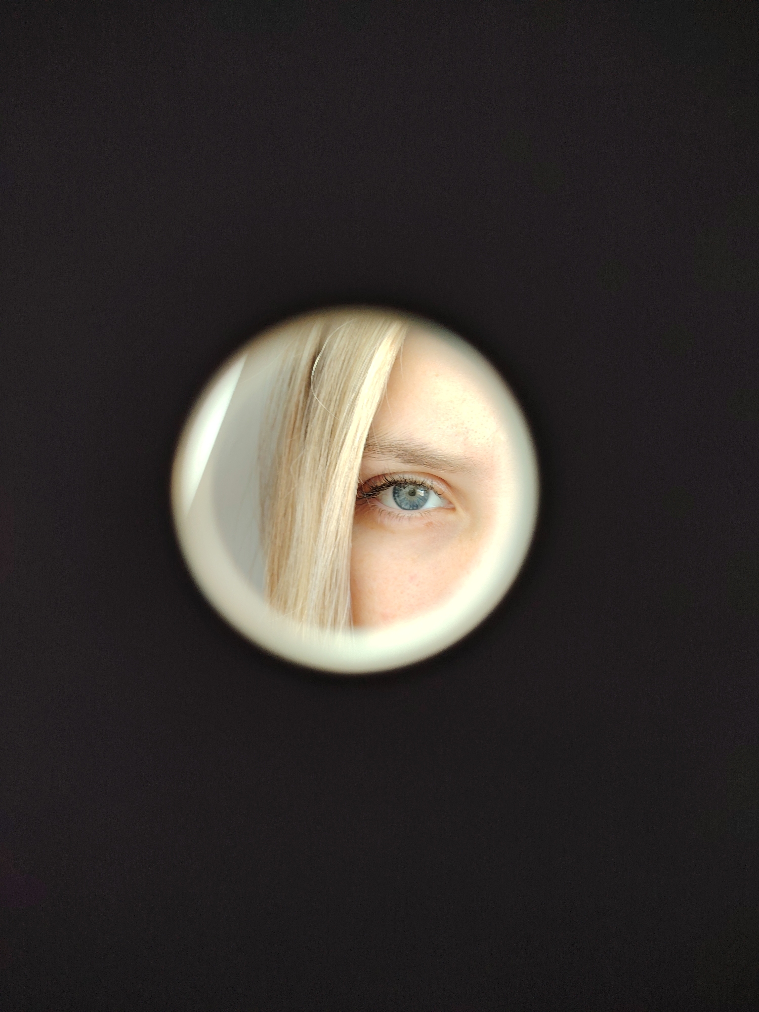 A close-up, centered, and vertical composition shows a single eye framed by blonde hair, viewed through a circular opening in a dark surface. The eye is light blue with visible eyelashes and eyebrow. The blonde hair partially obscures the face on the left side of the opening. The background surrounding the circular frame is a uniform, dark color. The image does not provide enough information to determine the location in Chaikovsky, Russia.