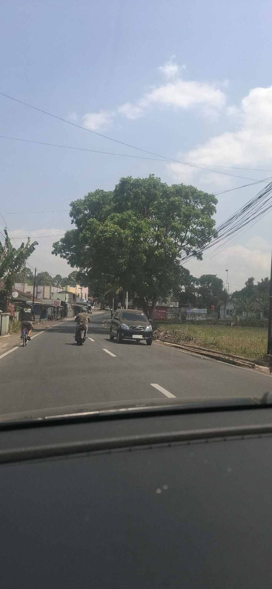 In the city of Cilaku, Indonesia, this image captures a view from inside a vehicle, looking out at a road scene. A dark-colored sedan is traveling on the right side of the asphalt road, which is marked with white dashed lines. In front of the sedan, a motorcycle is also heading in the same direction, with a rider wearing a patterned shirt. To the left of the road, a cyclist on a bicycle is moving away from the viewer. The road is lined with buildings and stalls on the left side, and a large, leafy green tree stands prominently in the center of the frame, partially obscuring the sky. Beyond the tree, more buildings and vegetation are visible. Overhead, power lines stretch across the blue sky, which has scattered white clouds. The overall environment suggests a semi-urban or rural setting in Indonesia.