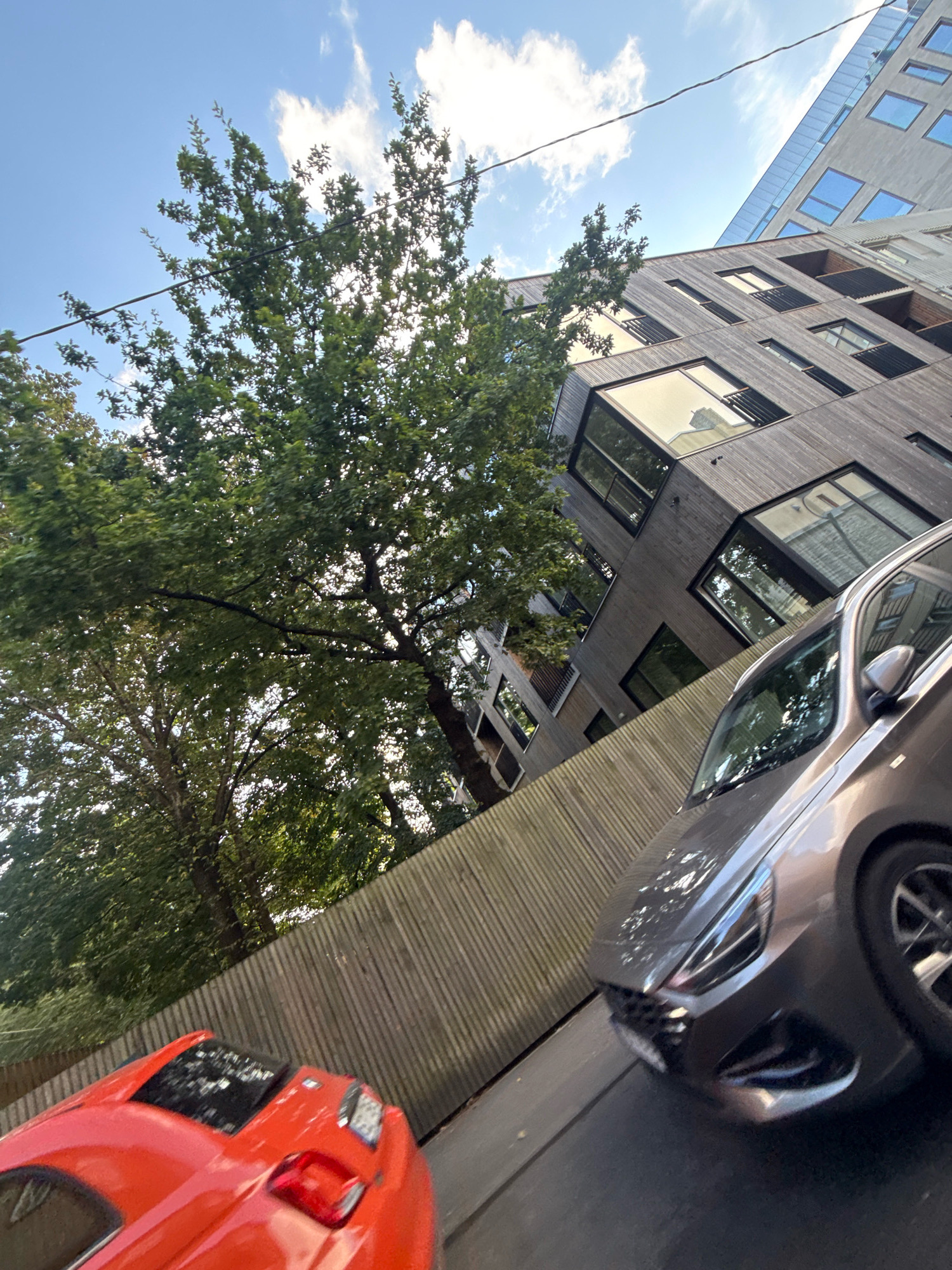 In Tallinn, Estonia, on a clear day with scattered clouds, a modern wooden-clad building with numerous large windows is visible, angled upwards. Trees with dense green foliage surround the building. In the foreground, a gray car is parked on a street, with its front end angled towards the viewer. To the left of the gray car, a portion of a bright red car is also visible. A wooden fence runs horizontally across the middle ground, separating the street from a grassy area. Power lines are strung across the sky.