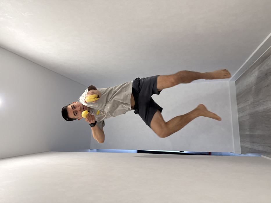An adult male subject is depicted mid-air in a narrow interior hallway, approximately centered in the frame. He is East Asian, with short dark hair, wearing a light grey t-shirt, dark grey shorts, and is barefoot. A black smartwatch is visible on his left wrist. Both arms are extended forward, with a ripe yellow banana held in each hand, pointed outwards like mock firearms. The banana in his right hand is extended further than the one in his left. His facial expression is concentrated. The hallway features smooth, light grey walls with a subtle texture, light grey wood-look laminate flooring, and white baseboards. A doorway is partially visible on the left side in the background. A single light source is present on the white ceiling directly above. The scene is located in a residential or commercial interior in Kazan, Russia.