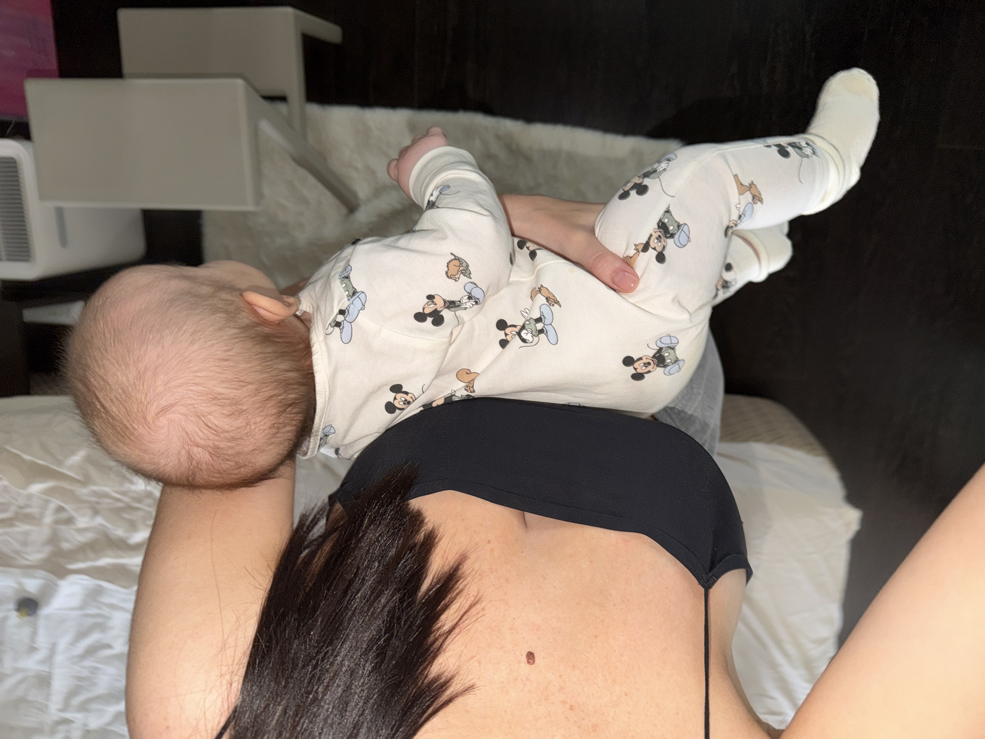 The image shows a baby lying on an adult, presumably its mother. The baby is lying on their back, wearing a white outfit with Mickey Mouse designs, and white socks. The baby's head is turned to the left. The adult is wearing a black bra or top. Some dark hair is visible, obscuring part of the adult's chest. The background includes a white bed, some white furniture, and dark flooring. The image location cannot be confirmed based on this image.