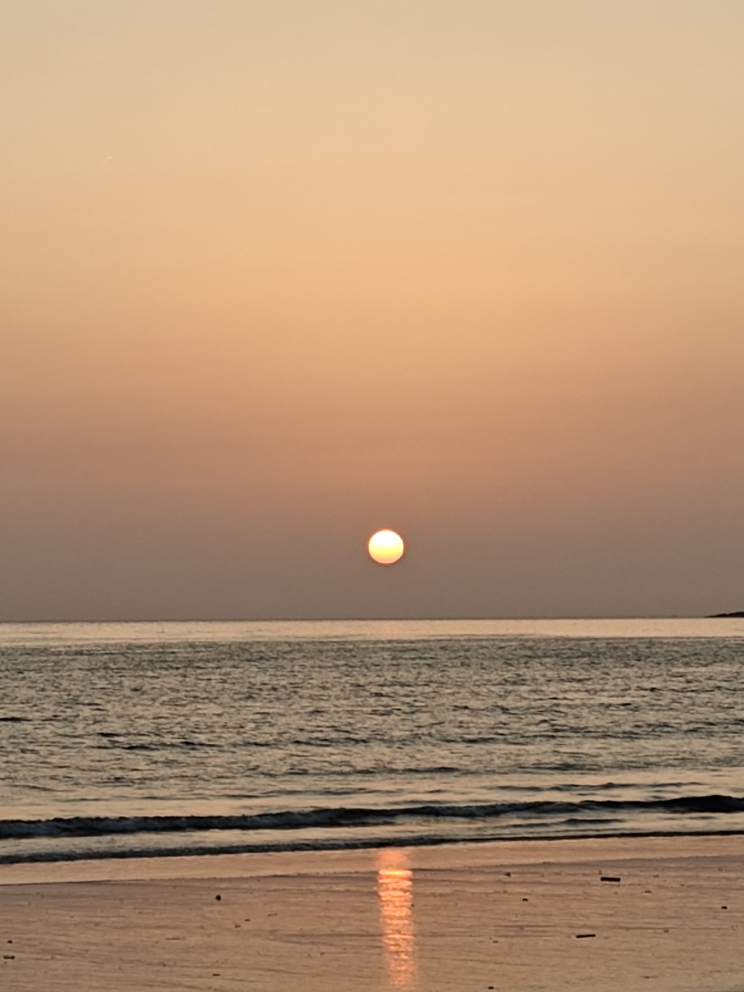 **Image Description**
**Location:** Mumbai, India
**Environment:** The image depicts a serene coastal scene during sunset.
**Sky:** A gradient of orange and peach hues fills the sky, indicating either sunrise or sunset.
**Water:** A calm sea stretches to the horizon, reflecting the warm colors of the sky. Gentle waves meet the shore.
**Beach:** The foreground consists of a sandy beach, partially wet from the receding tide. The wet sand reflects the sunlight, creating a mirrored effect.
**Sun:** A soft orange sun is visible near the horizon, casting a golden glow across the scene.
**Objects:** In the distance, a small landmass is visible on the right side of the picture.