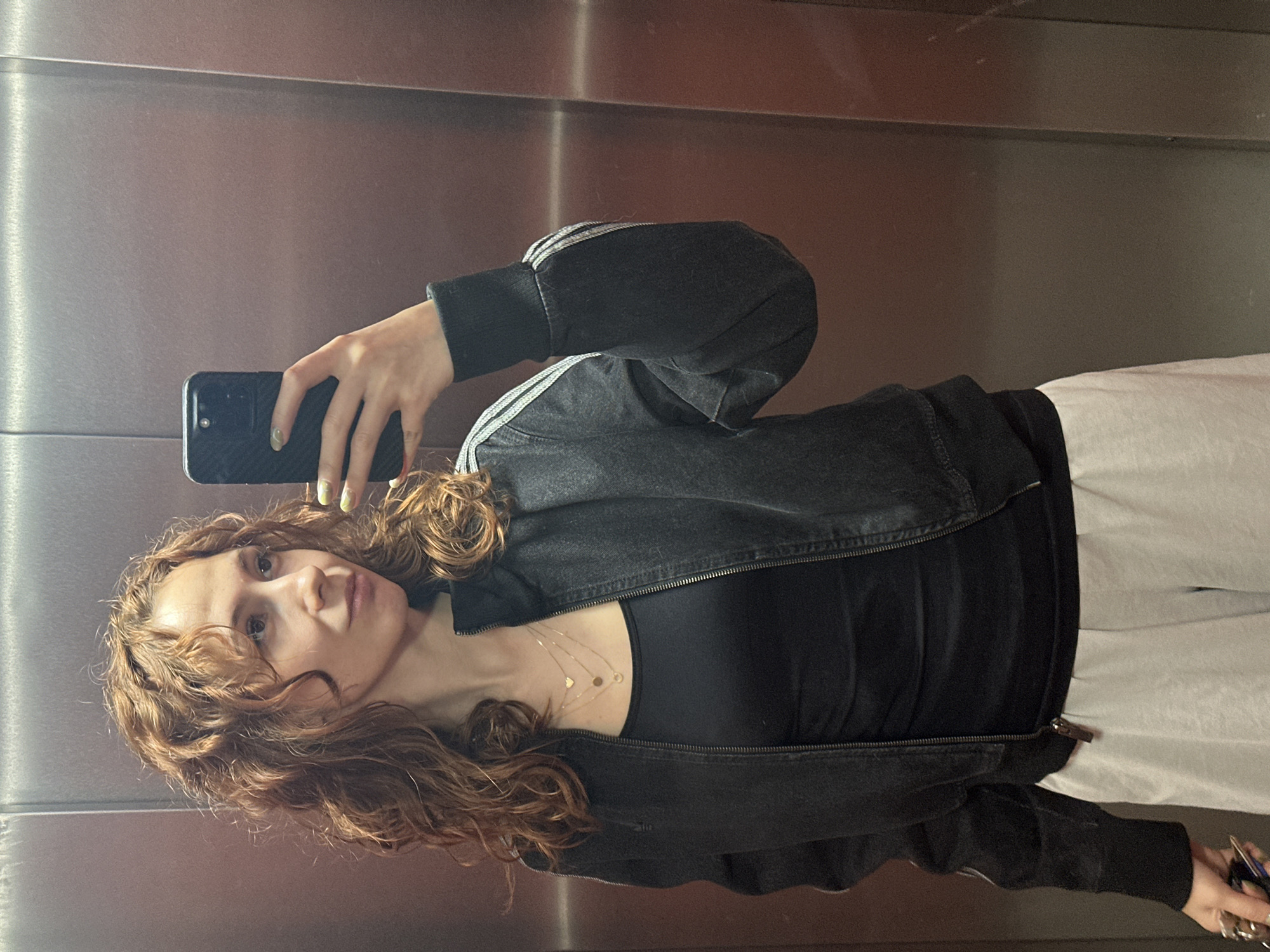A young woman is taking a selfie in an elevator. She has curly auburn hair and is wearing a black tank top and a black Adidas zip-up jacket with white stripes on the sleeves. She is also wearing a delicate gold necklace. She is holding a black smartphone in her left hand, and her right hand is visible near the bottom right corner of the image, holding keys. She appears to be standing in front of a brushed steel elevator wall. The location in the city of Novi Sad, Serbia, cannot be determined from the visual information in the image.