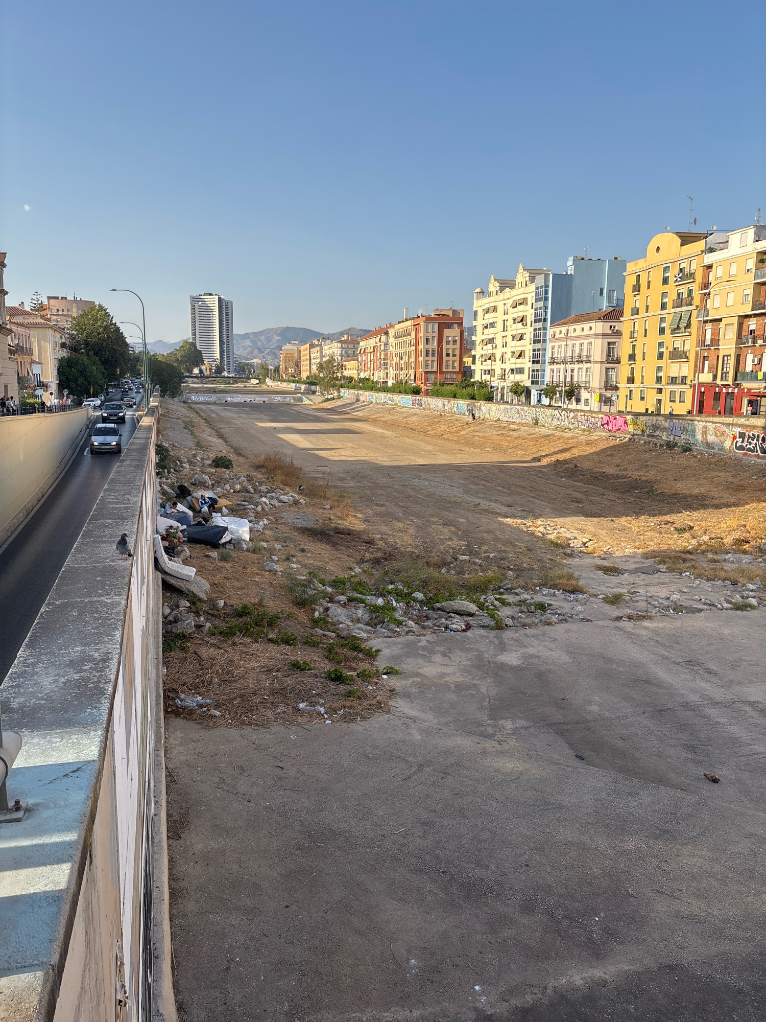 The image depicts a dry riverbed in Málaga, Spain, with a road and buildings on the left. The riverbed is mostly barren earth with some dry vegetation and scattered debris. Along the retaining wall of the riverbed, graffiti is visible. In the foreground, on the left side, a concrete wall runs alongside the road. A pigeon is perched on this wall, and a pile of discarded mattresses and personal belongings sits on the ground near the riverbed's edge, suggesting the presence of homeless individuals. On the right side of the image, the riverbed slopes down, showing more debris and dry earth. In the background, a row of colorful buildings lines the opposite side of the riverbed, and beyond them, a modern tall building and mountains are visible under a clear blue sky. The sun is casting long shadows, indicating it is likely late afternoon or early evening.