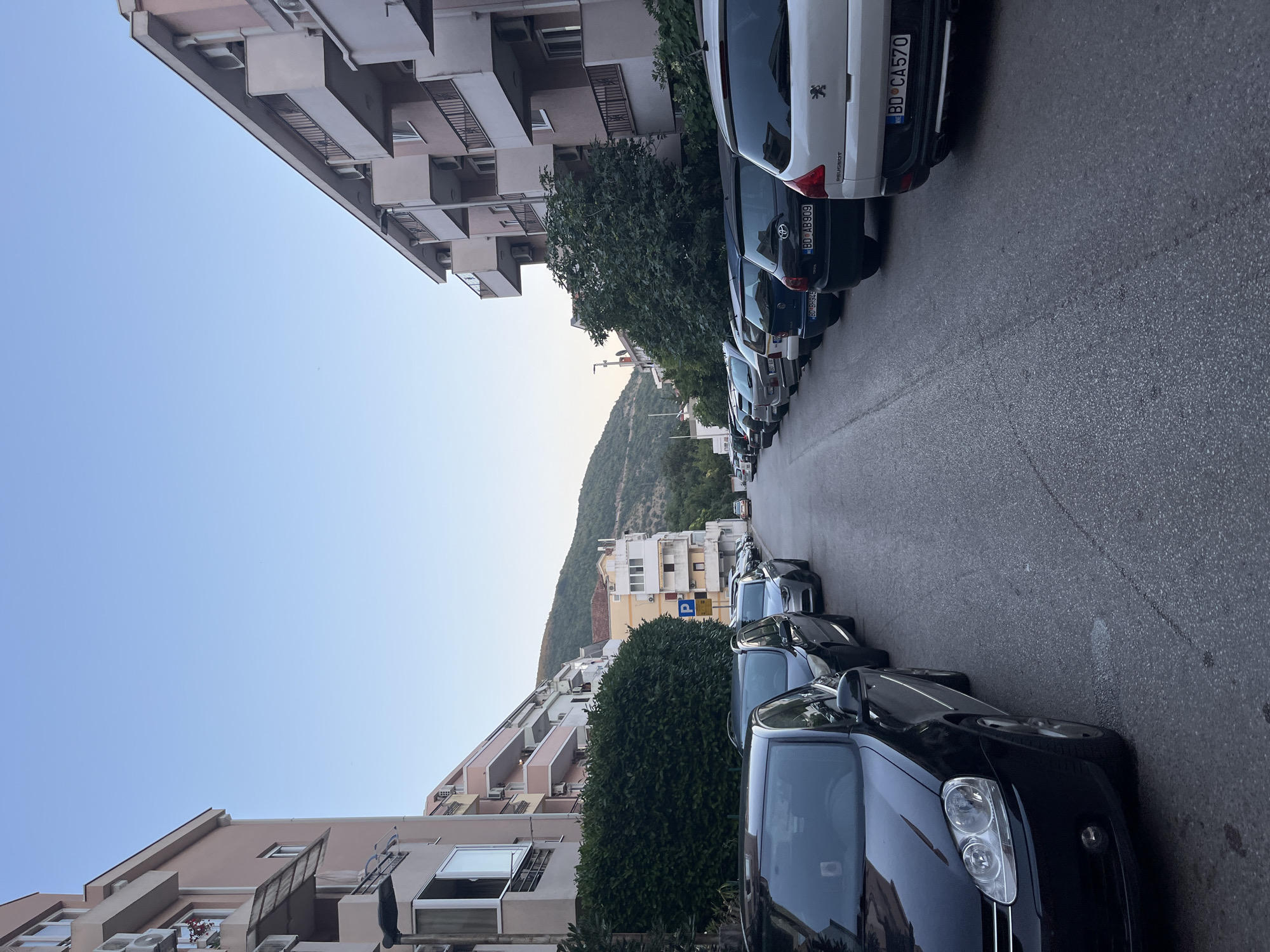 A street in Budva, Montenegro, is lined with parked cars on both sides, with apartment buildings on either side of the street. The buildings are multi-story with balconies. A lush green hedge runs along the left side of the street in front of one of the apartment buildings. The sky is clear and blue. The road is asphalt. A parking sign is visible on the right side of the street. The scene appears to be in a residential area.