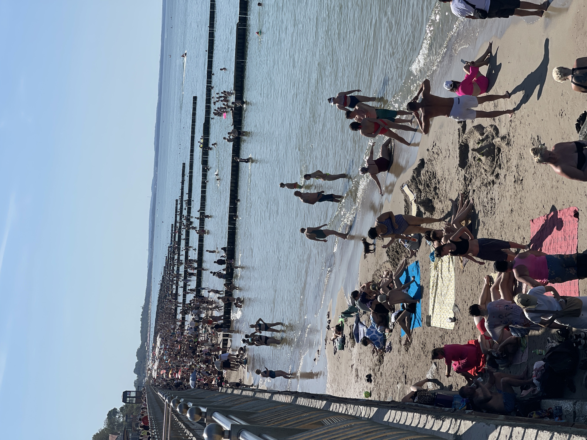The image shows a crowded beach in Zelenogradsk, Russia, on a sunny day. Numerous people are sunbathing, swimming, and walking along the shore. The water is calm, with wooden structures extending into the sea, possibly as breakwaters. A walkway with a railing runs along the left side of the image, separating the beach from a treed area and buildings in the distance. People are scattered across the sandy beach, some under umbrellas, while others are directly exposed to the sun. The sea extends to the horizon, meeting a clear blue sky.