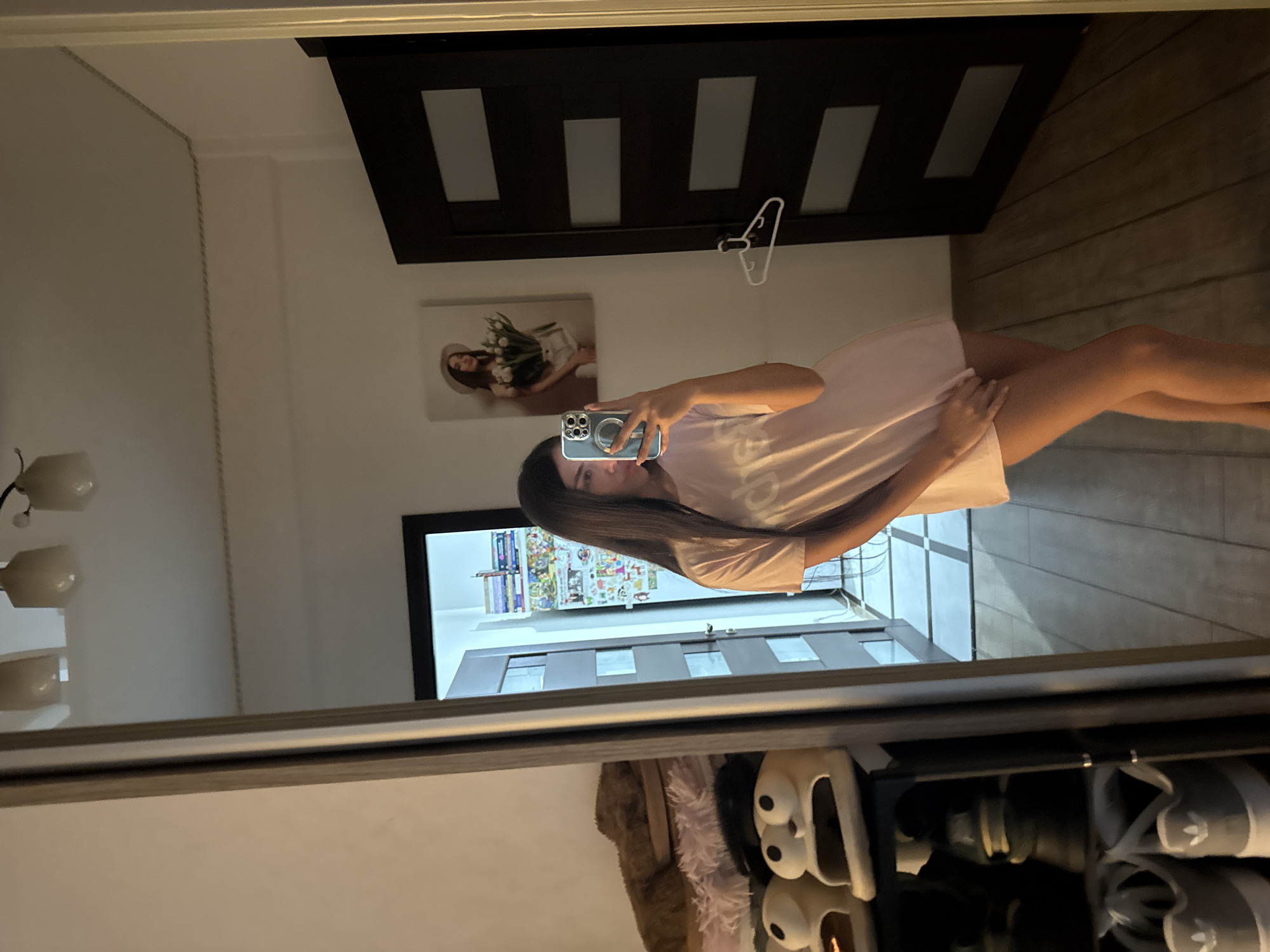 A young woman is taking a mirror selfie in a room. She is wearing a pink t-shirt dress and has long dark hair. In the reflection of the mirror, a shoe rack with several pairs of shoes is visible to the left. Behind the woman, a painting of a woman holding flowers hangs on the wall, and a dark door with glass panels is to her right. The room has wooden flooring and a tiled floor visible through an open doorway in the background. The scene appears to be within a residential setting in Chișinău, Moldova.