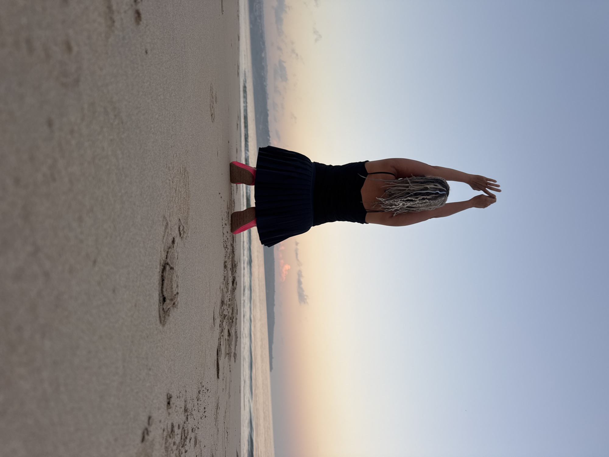 An overhead shot shows a person on a beach in Kuta, Indonesia. The person is facing away from the camera, with their arms raised in the air. They are wearing a black tank top and a dark pleated skirt. Pink shoes are visible on their feet. The beach is made of sand and the ocean is in the background. The sky is a pale blue with hints of pink and orange from a sunrise or sunset.