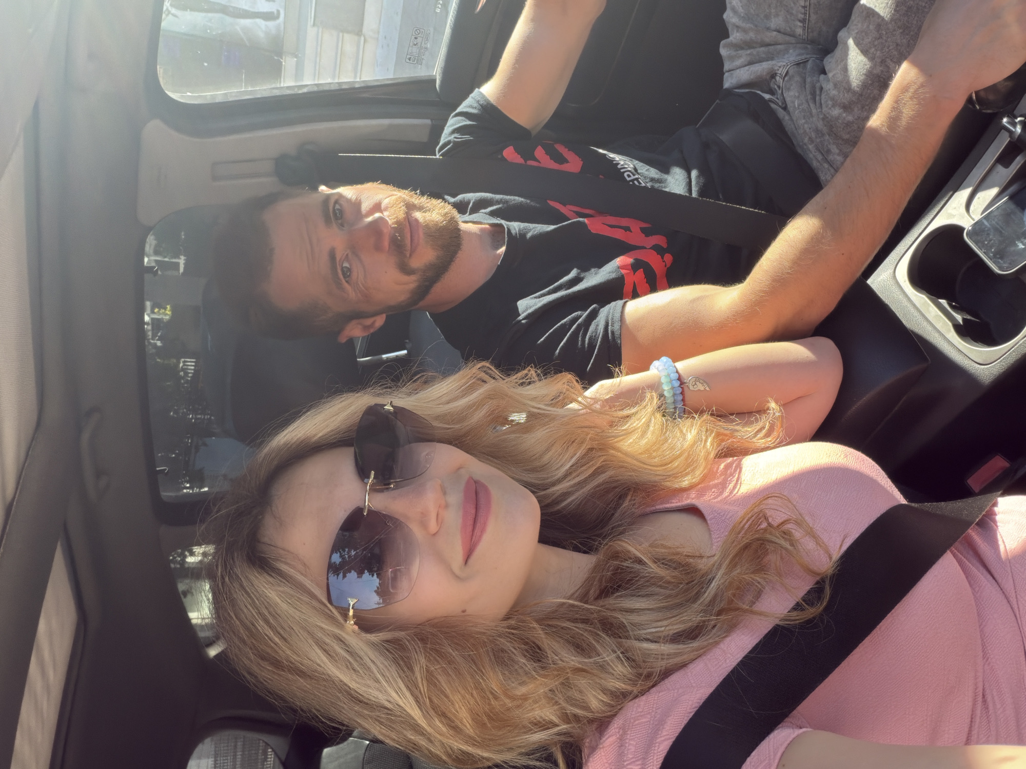 The image shows two individuals, a man and a woman, seated inside a car during bright daytime. The composition suggests a selfie or a photo taken from the perspective of the woman, who is in the foreground.
The woman has long, wavy blonde or light brown hair and is wearing a pink top. She is looking directly at the camera with a subtle smile and is wearing large, dark aviator-style sunglasses. Her left arm is visible, adorned with several light-colored beaded bracelets, one featuring a small metallic charm. A black seatbelt is visible across her chest.
In the background, the man is also looking towards the camera with a neutral expression, sporting a beard. He is wearing a black t-shirt with stylized red text, where the word "KEEPING" is clearly visible, and part of another word follows, possibly "IT REAL". He appears to be wearing grey shorts or trousers and also has a black seatbelt fastened. His right arm is resting, with his forearm visible.
The interior of the car is visible around them, including parts of the dashboard, seats, and a center console with cup holders. Bright sunlight streams into the car, causing some lens flare in the upper left corner and highlighting the subjects. Reflections in the car windows and the woman's sunglasses show blurry outdoor scenery, possibly buildings and trees, consistent with being in a sunny location like Budva, Montenegro. The overall atmosphere is relaxed.