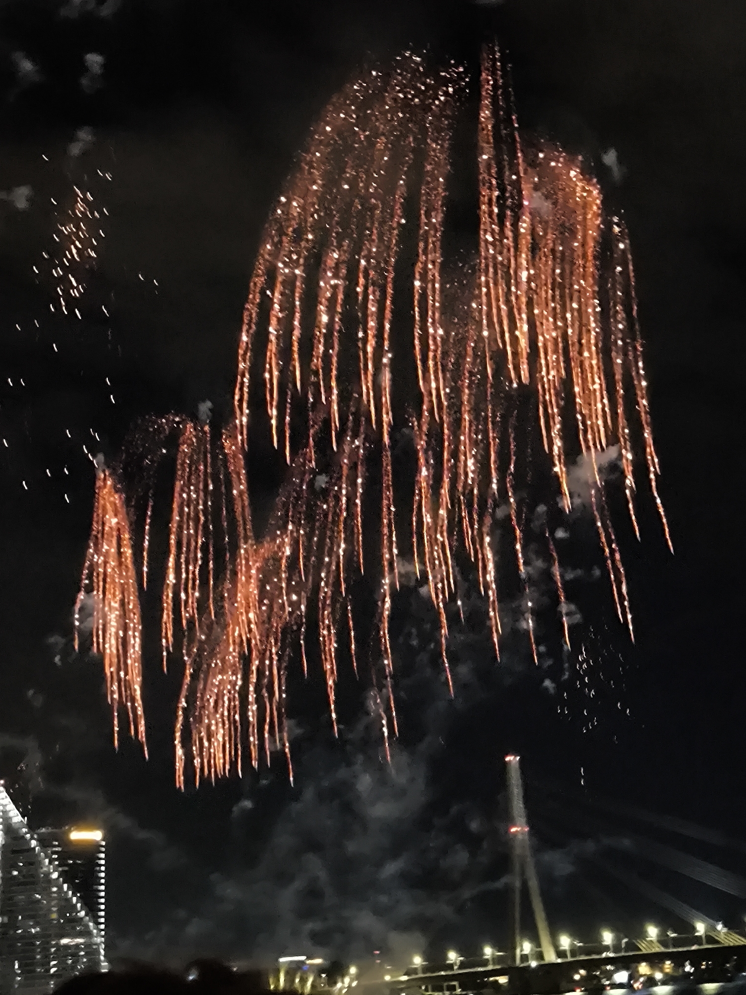 The image captures a nighttime fireworks display over a city, likely Riga, Latvia, given the distinctive bridge structure. The sky is dark, illuminated by streaks of orange and red fireworks cascading downwards, creating a curtain-like effect. Below the fireworks, a modern bridge with illuminated pylons stretches across the scene, its cables faintly visible. In the lower left, a tall, illuminated building with a geometric facade is partially visible. The scene suggests a festive event, with people present at the bottom of the frame, their silhouettes suggesting a crowd gathered to watch the spectacle. The overall atmosphere is one of celebration and wonder, with the vibrant fireworks contrasting against the dark night sky and the urban landscape.
