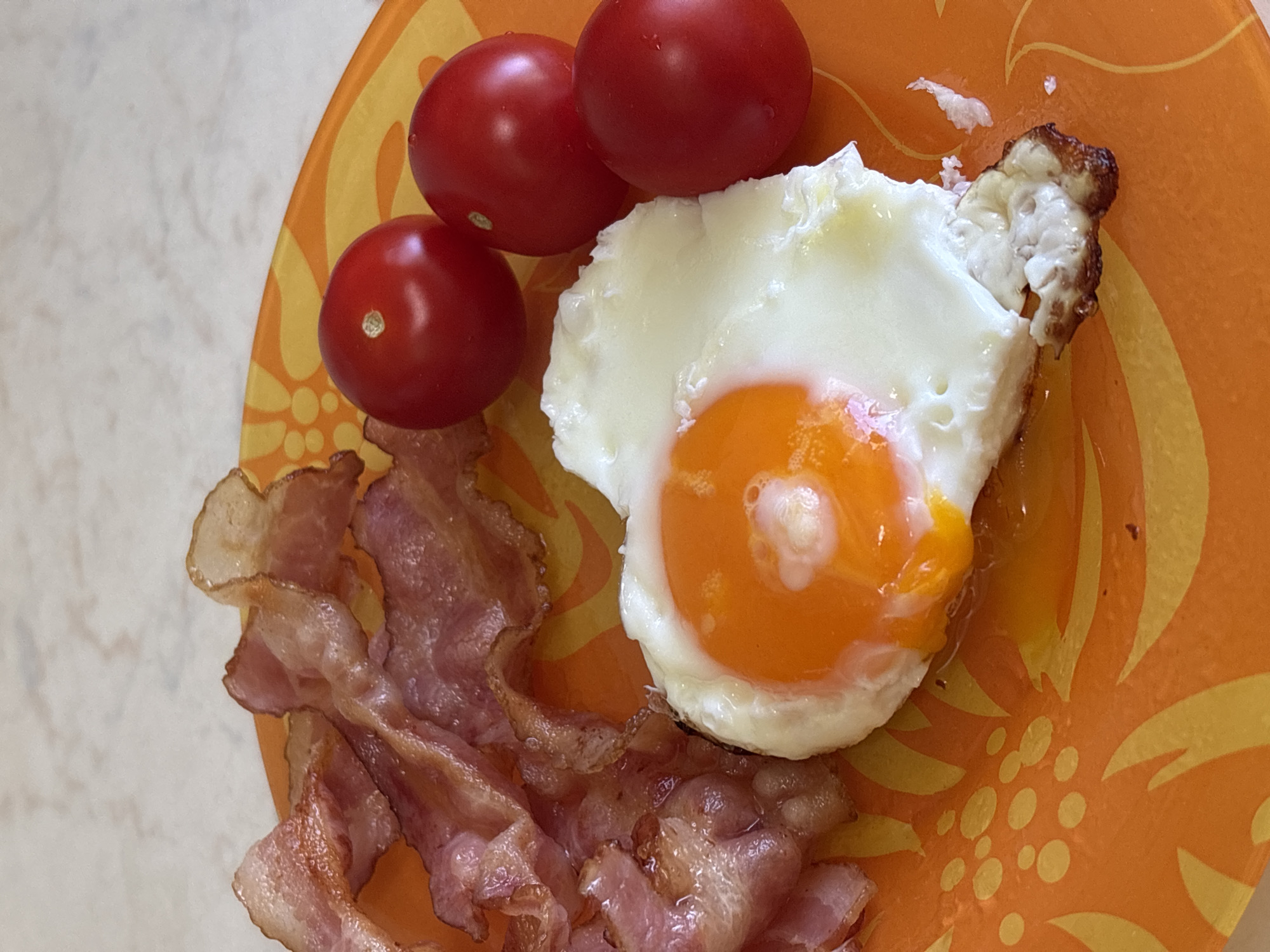 On an orange floral plate are three red cherry tomatoes, a fried egg with a runny yolk, and several strips of cooked bacon. The plate sits on a surface with a light, textured pattern. The scene appears to be a breakfast or brunch setting. There is no indication of a location in Rusanovo, Russia.