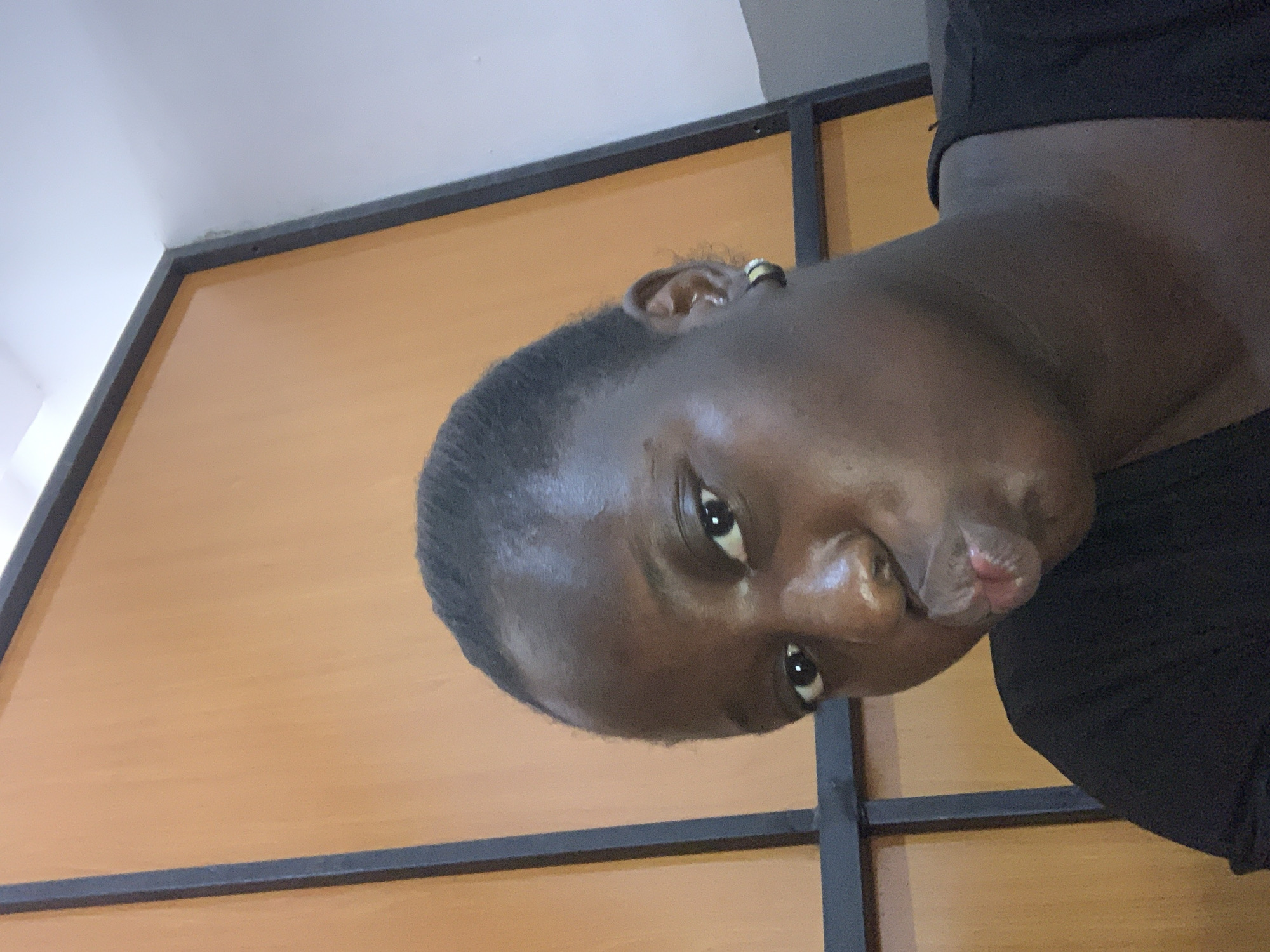The image is a close-up, selfie-style photograph of a young Black woman with dark skin and black hair pulled back. She is wearing a black shirt and a small hoop earring in her visible left ear. Her lips are pursed, and her head is tilted slightly. The background consists of a wooden panel with a dark frame, suggesting an indoor setting. The lighting is somewhat diffuse, and the image quality is adequate for facial recognition. The location is not discernible from the image.