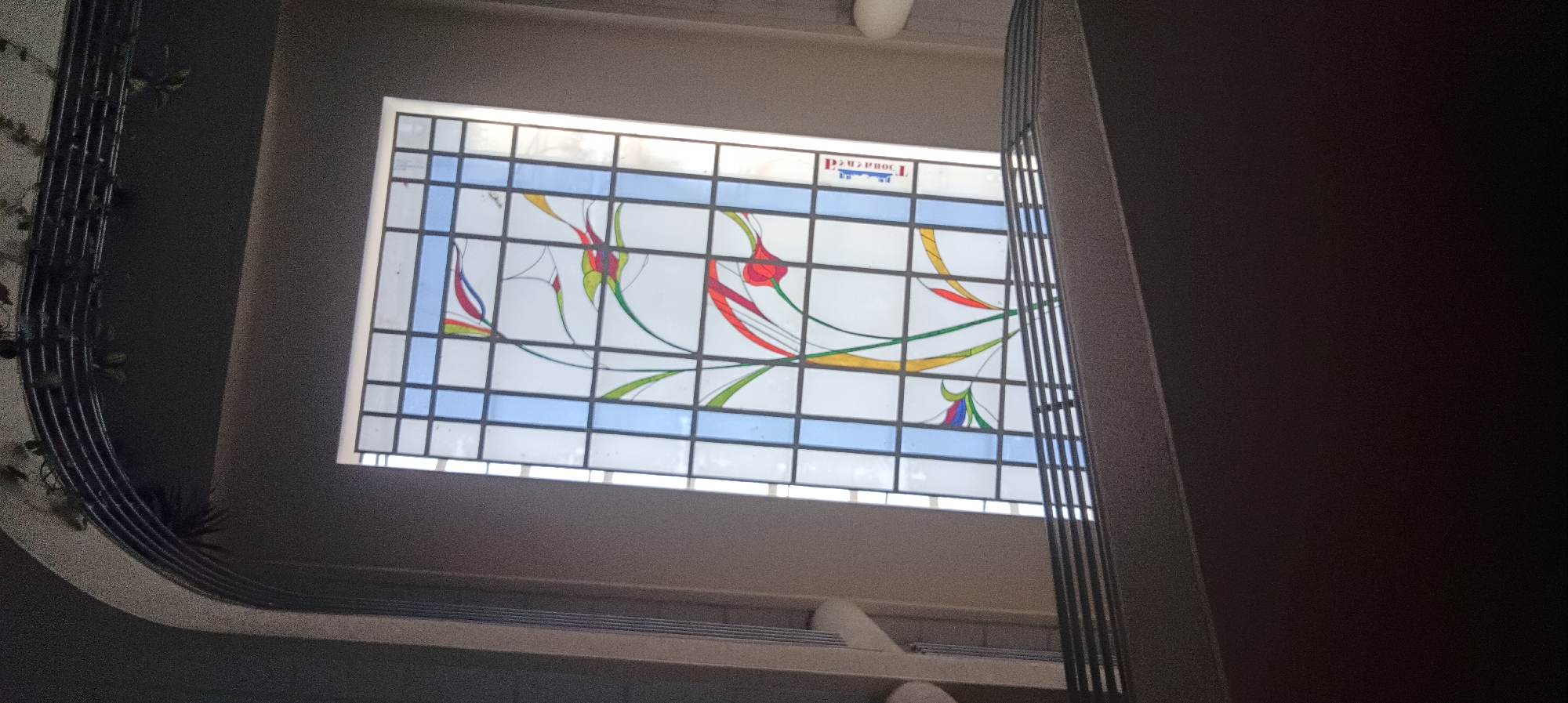 The image shows a stained glass window in what appears to be a stairwell or hallway. The window is rectangular and features a geometric design with abstract floral elements in red, yellow, green, and blue. The glass is set within a grid of dark lead or metal framing. Above the stained glass, there is a smaller pane of glass with some text, possibly a logo or inscription. To the left of the window, a dark, curved structure with thin vertical supports is visible, possibly part of a railing or decorative element, with some greenery attached. The walls surrounding the window are a neutral, light brown color. The lighting suggests the window is illuminated from behind, casting a glow. This architectural feature is located within the Faculty of Technical Sciences at the University of Novi Sad, Serbia, specifically in the section of the building that houses the Department of Architecture and Urban Planning.