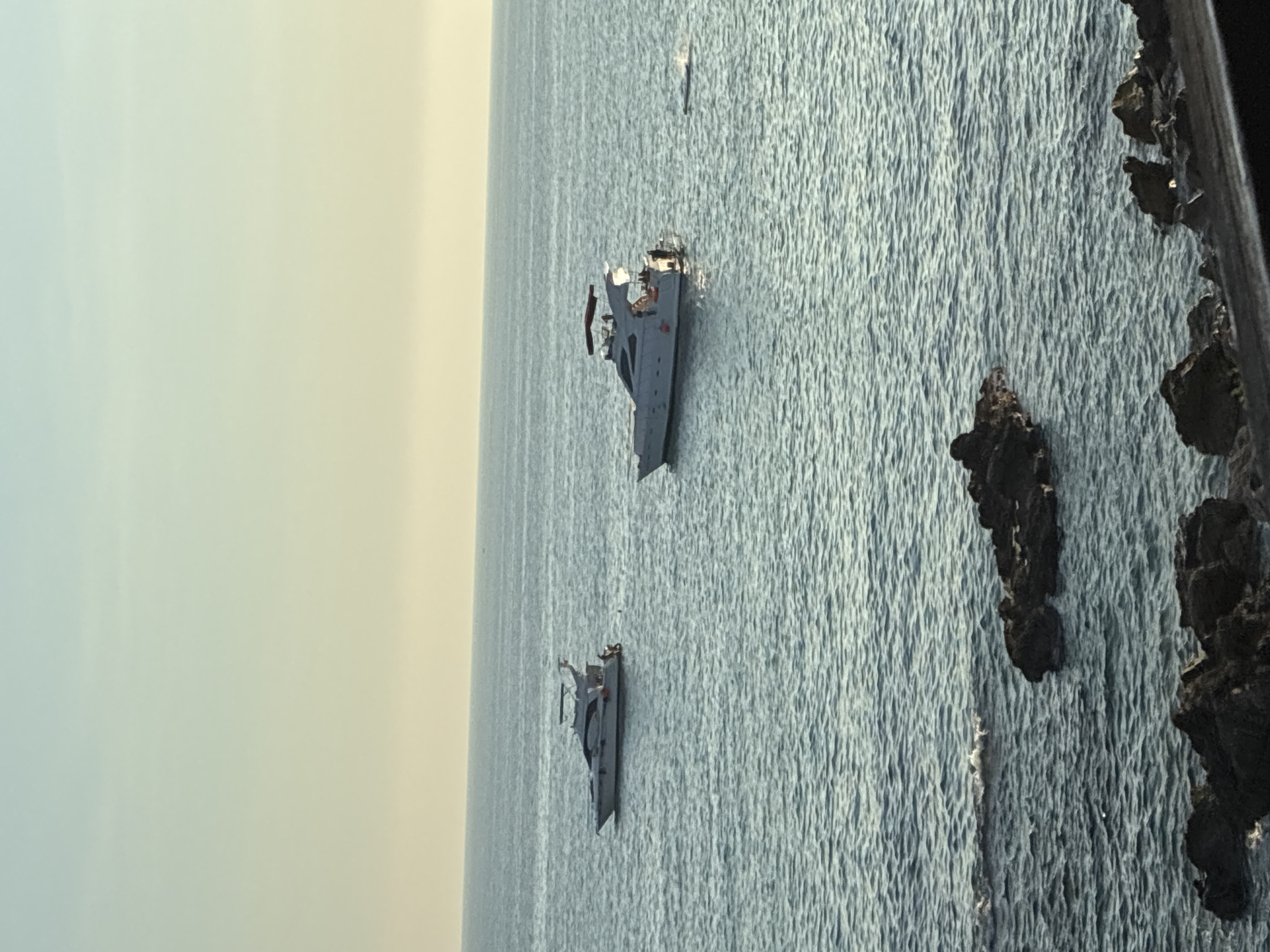 Two yachts are anchored in the calm sea, with the coast of Alanya, Turkey visible in the foreground. The larger yacht, with a white hull and a dark bimini top, is positioned to the right of the frame. Several people are visible on its deck. A smaller yacht with a white hull is to the left. Dark, rocky outcrops break the surface of the water in the foreground, suggesting a shallow area near the shore. The sky is a clear, pale blue, transitioning to a yellowish hue at the horizon, indicating either early morning or late afternoon.