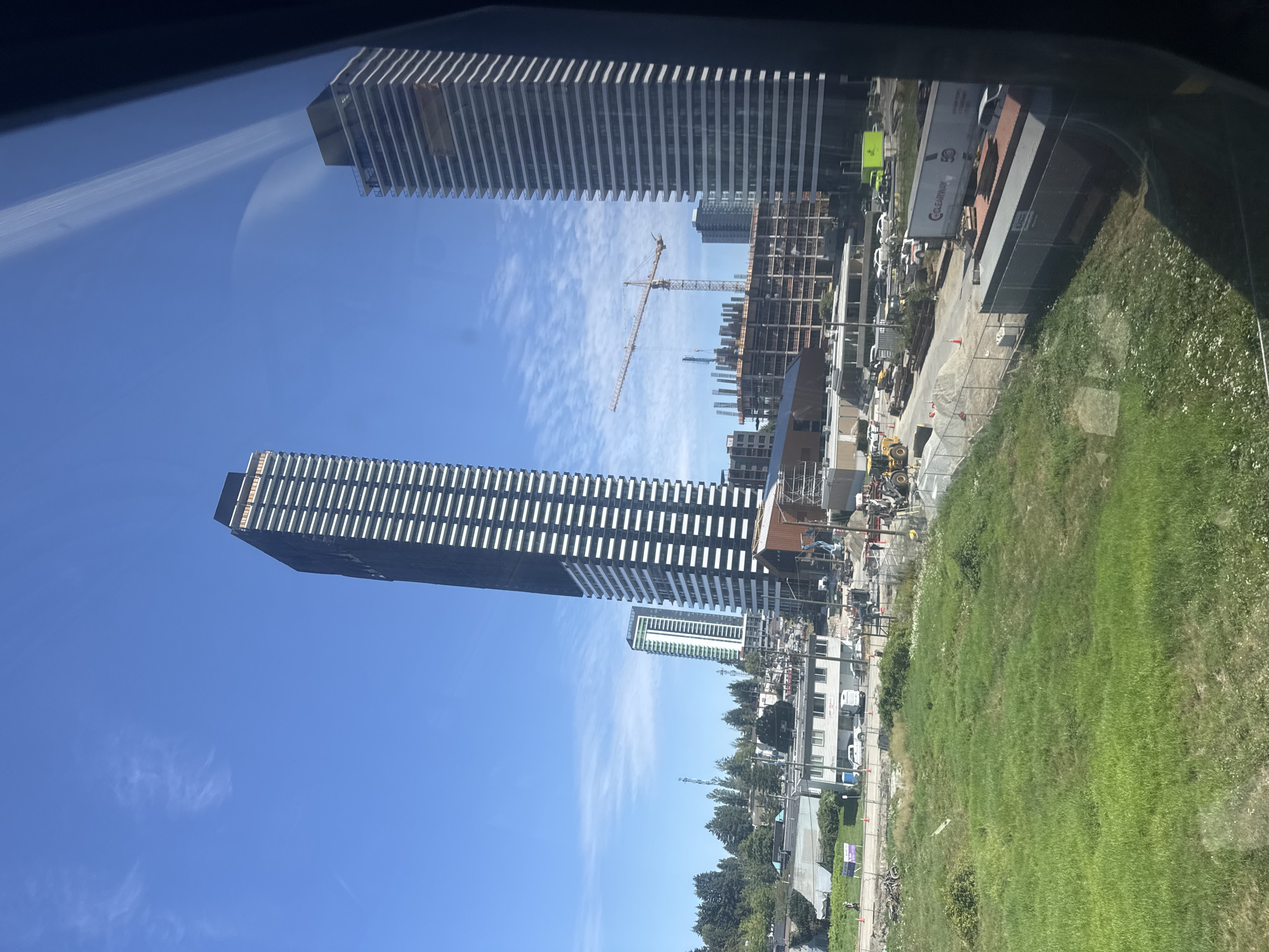 This image captures a city skyline in Surrey, Canada, featuring several modern high-rise buildings under construction. The foreground is dominated by a large, untamed green grassy field, bordered by a chain-link fence. Beyond the field, a construction site is active, with scaffolding, building materials, and construction vehicles visible. A prominent feature is a tall, dark-colored skyscraper with a white grid pattern, appearing to be nearing completion. To its right, another tall, white-and-glass building stands, also likely residential or office space. In the background, a construction crane is operational, indicating ongoing development. The sky is a clear, bright blue with scattered white clouds, suggesting a sunny day. The reflection of the sky and buildings is visible on what appears to be a window, implying the photo was taken from inside a vehicle or building.