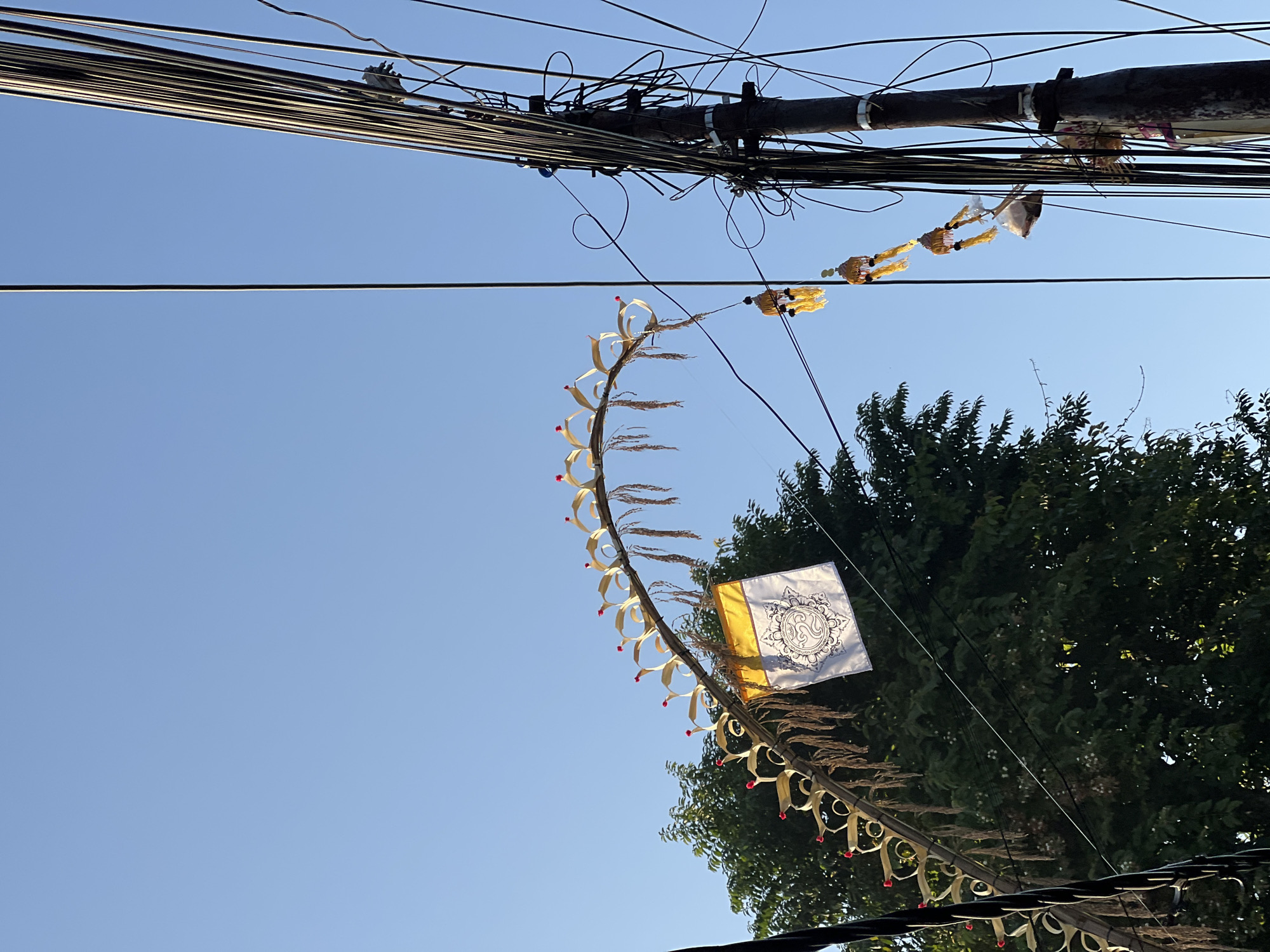 In Kuta, Indonesia, a clear blue sky serves as the backdrop for a street scene featuring overhead utility wires and decorative elements. A large, curved bamboo structure, adorned with woven leaf-like decorations tipped with red accents and strands of dried rice, spans across the frame. A white flag with yellow trim, featuring a black circular emblem, hangs from the center of this structure. To the right, a utility pole is heavily laden with numerous black wires, some of which are neatly bundled while others form a tangled network. Suspended from these wires are three small, ornamental yellow and gold objects, likely decorative tassels or small banners. In the background, a dense green tree provides a contrasting natural element to the urban infrastructure. The lighting suggests it is daytime, with the sun casting shadows and illuminating the scene from the side.