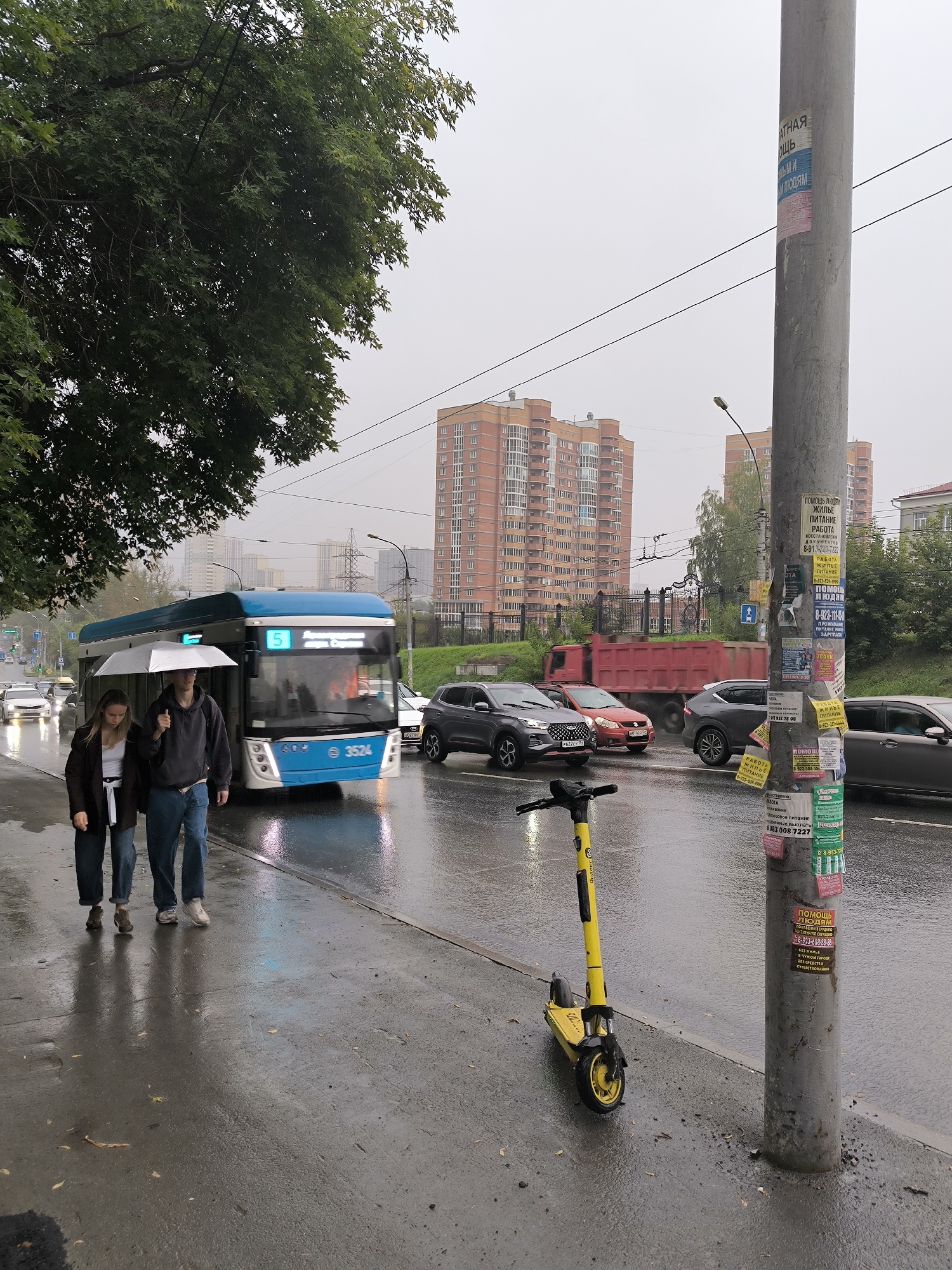 In Novosibirsk, Russia, on a rainy day, a young couple walks under a shared white umbrella on a wet sidewalk next to a street. The man, wearing a dark hoodie and jeans, holds the umbrella over himself and the woman, who is in a dark jacket and jeans. A blue and white trolleybus, numbered '5' and with license plate '3524', is stopped nearby. A yellow electric scooter stands on the sidewalk in the foreground. In the background, several apartment buildings with orange facades rise against a grey, overcast sky. Traffic is visible on the wet road, including a red dump truck. A concrete lamppost covered in various advertisements stands on the right side of the frame.