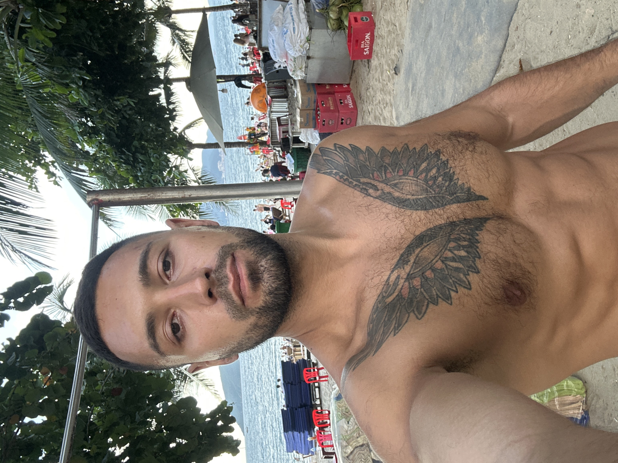 A man with a beard and a winged tattoo on his chest is taking a selfie on a beach. He is shirtless, revealing his muscular physique. In the background, there is a body of water, palm trees, umbrellas, beach chairs, and people. The scene appears to be a lively beach resort area in Nha Trang Ward, Vietnam.