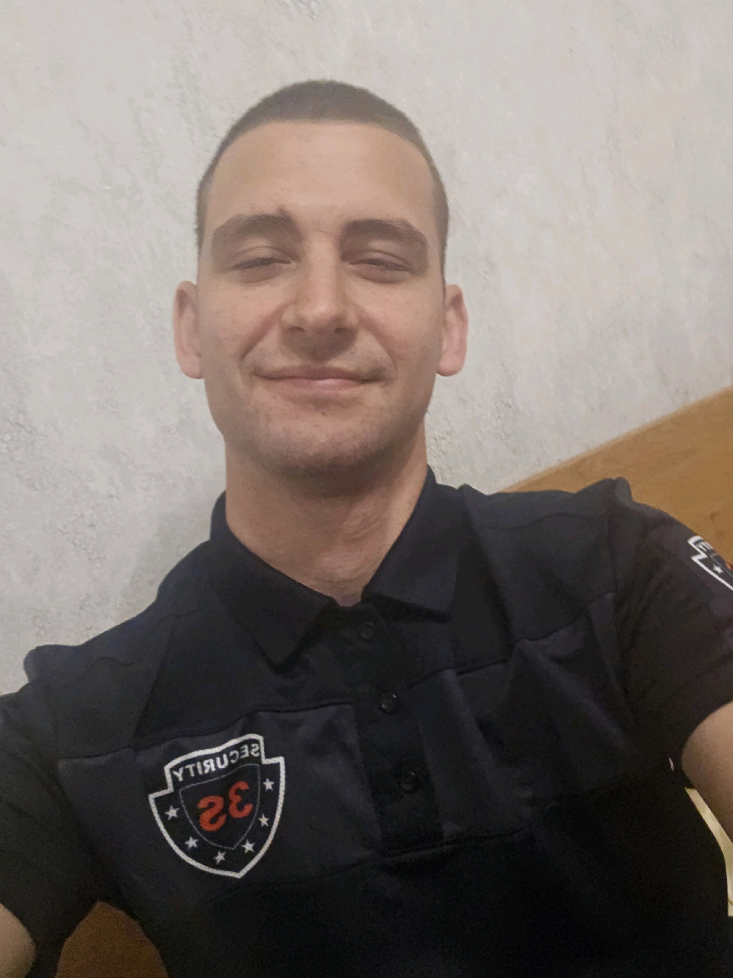 A man with short hair and a light complexion is photographed from the chest up, looking directly at the camera with a slight smile. He is wearing a dark blue polo shirt with a patch on the left chest area. The patch is a white shield shape with a black border. Inside the shield, "SECURITY" is written in black, reversed letters. A red number "32" is prominently displayed in the center of the shield, with smaller white stars surrounding it. The background consists of a textured light-colored wall on the left and a wooden surface on the right. The lighting is even, casting soft shadows. The man's location is not specified in the image or metadata, and there is no information available to determine if he is in Haskovo, Bulgaria.