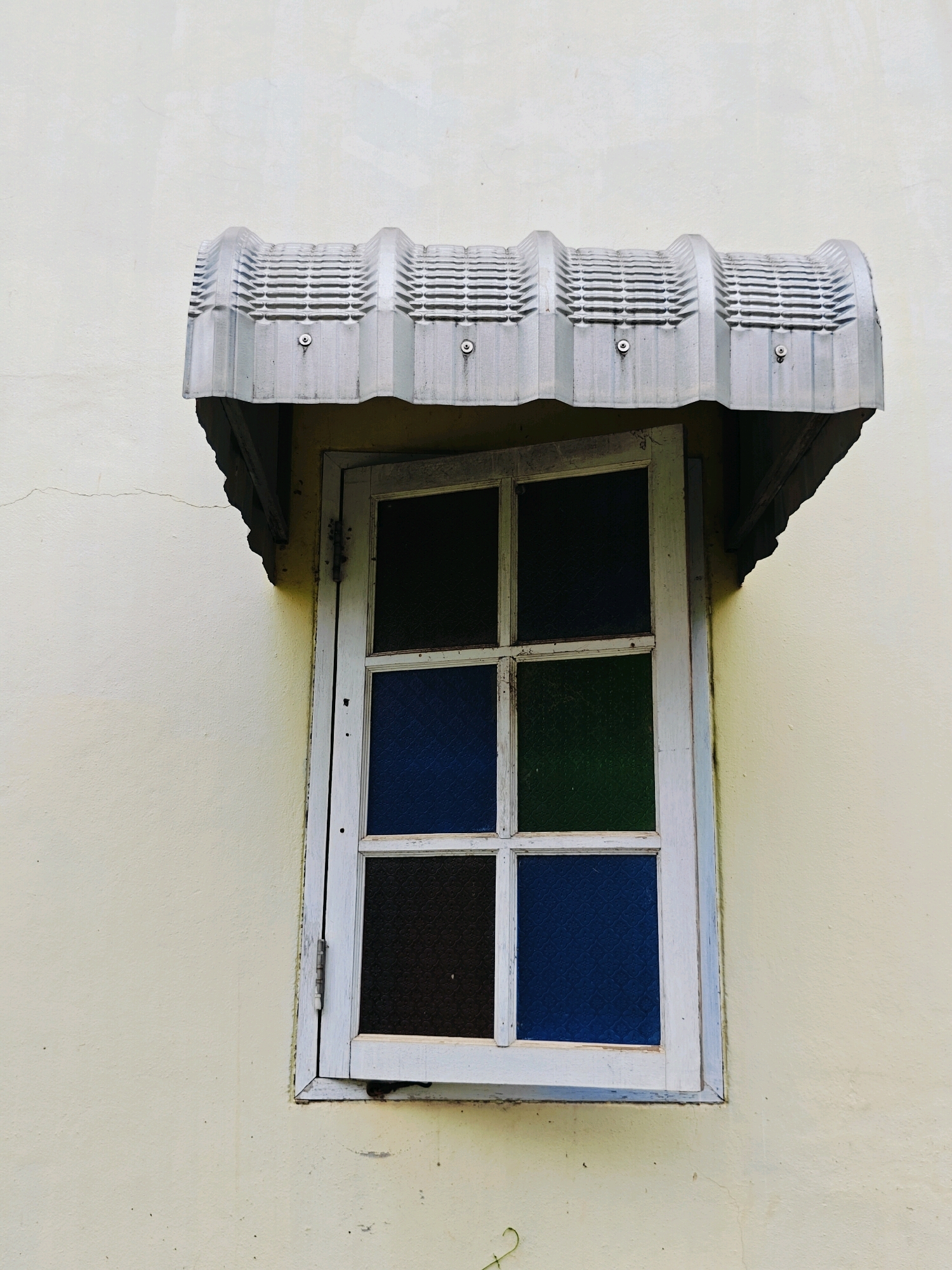 A weathered, off-white wall in Khao Yai, Thailand, serves as the backdrop for a simple, white-framed window. The window is divided into six panes, each filled with colored glass: two panes are dark green, two are deep blue, and two are dark brown or black. Above the window, a corrugated metal awning, also weathered and gray, is attached to the wall with visible screws. The awning has a decorative, wavy edge. The wall shows signs of age, with a few faint cracks and discoloration. The overall scene depicts a quiet, somewhat aged architectural detail within its environment.