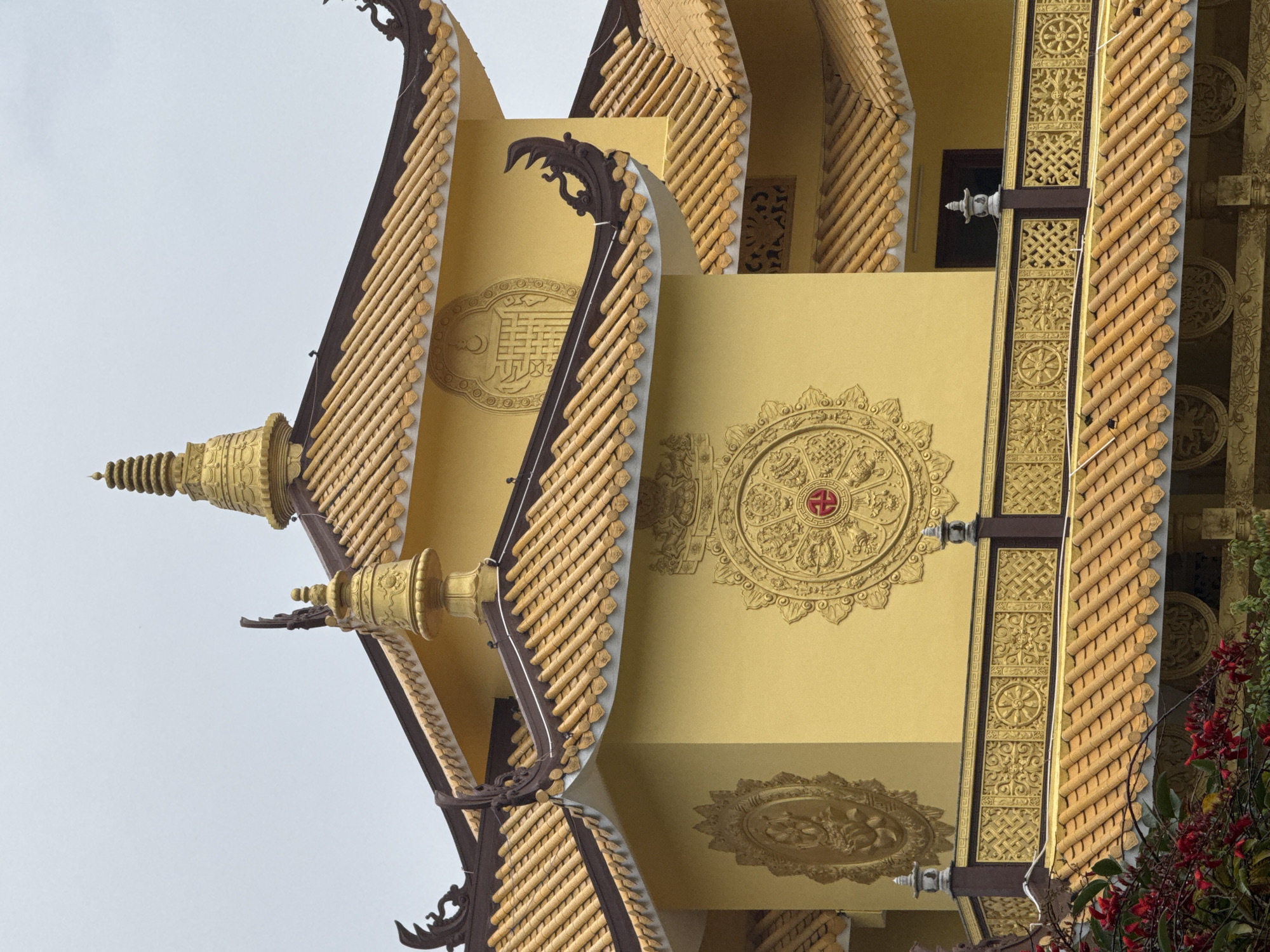 The image shows the golden, ornate roof and upper facade of a Buddhist temple. The architecture features multi-tiered, curved roofs covered in light brown tiles. The walls are a vibrant yellow and decorated with intricate golden circular emblems, some depicting lotus flowers and other Buddhist symbols. A decorative railing with geometric patterns and further circular emblems separates the upper levels. At the very top, a golden stupa-like structure rises against a pale, overcast sky. There are no people visible in the image. This architectural style is characteristic of Linh Phuoc Pagoda in Da Lat, Vietnam.