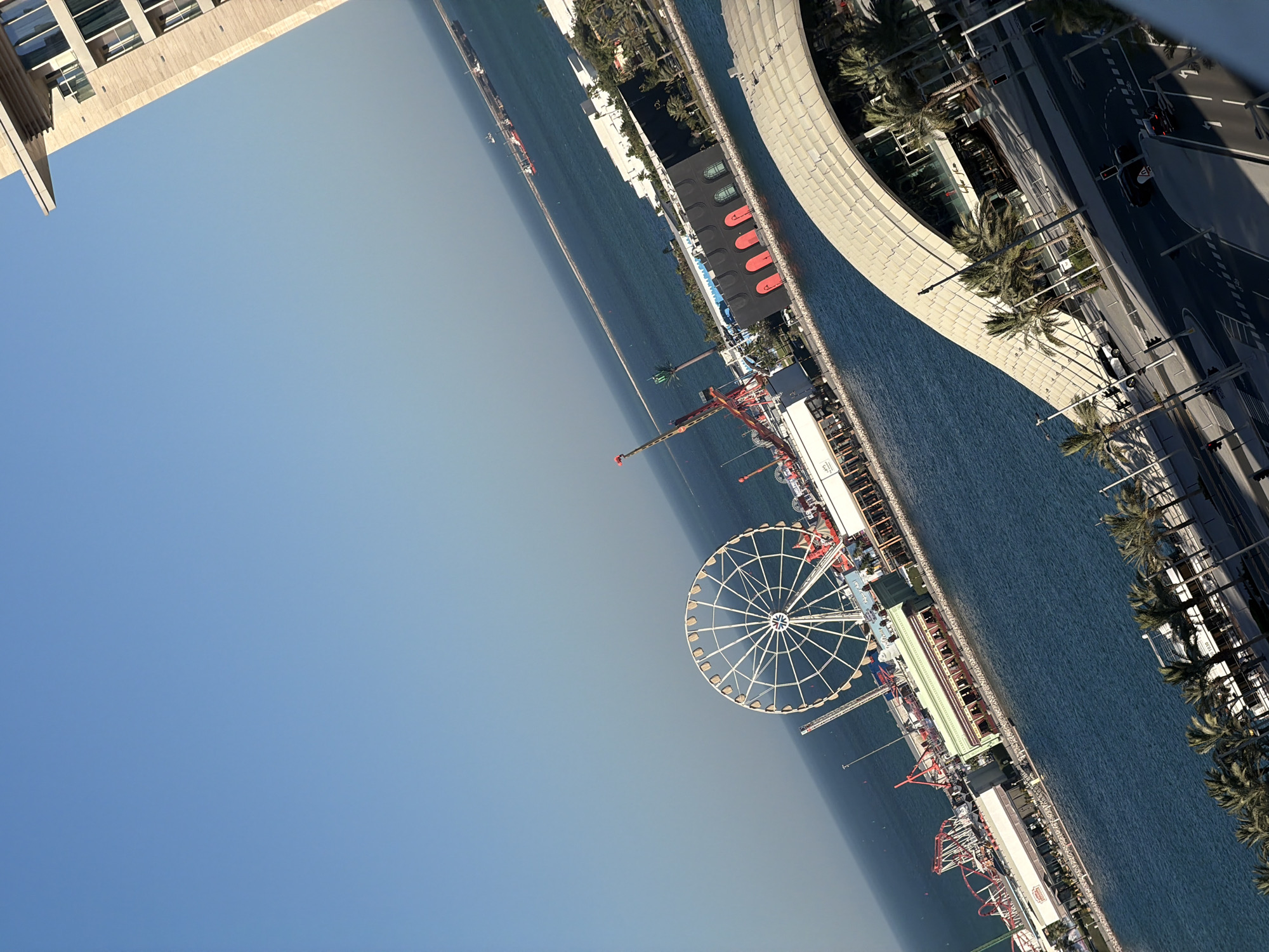 An eye-level, wide-angle shot captures a bright, sunny day in an unknown city. The sky is a clear, light blue, with a sliver of a modern building on the far right, featuring beige stone accents and large windows. In the middle ground, a large Ferris wheel dominates the scene, its white spokes clearly visible against the blue water. Surrounding the Ferris wheel is an amusement park with red and white roller coasters and other attractions. Buildings with black facades and arched red doorways are visible along the waterfront, interspersed with palm trees. A long pier extends into the calm, blue ocean, with a few cargo ships dotted in the distance. In the foreground, a curved, beige-roofed modern structure partially obscures a street below. Cars are seen driving on the street, and palm trees line the sidewalks. The water is calm and reflects the clear blue sky.