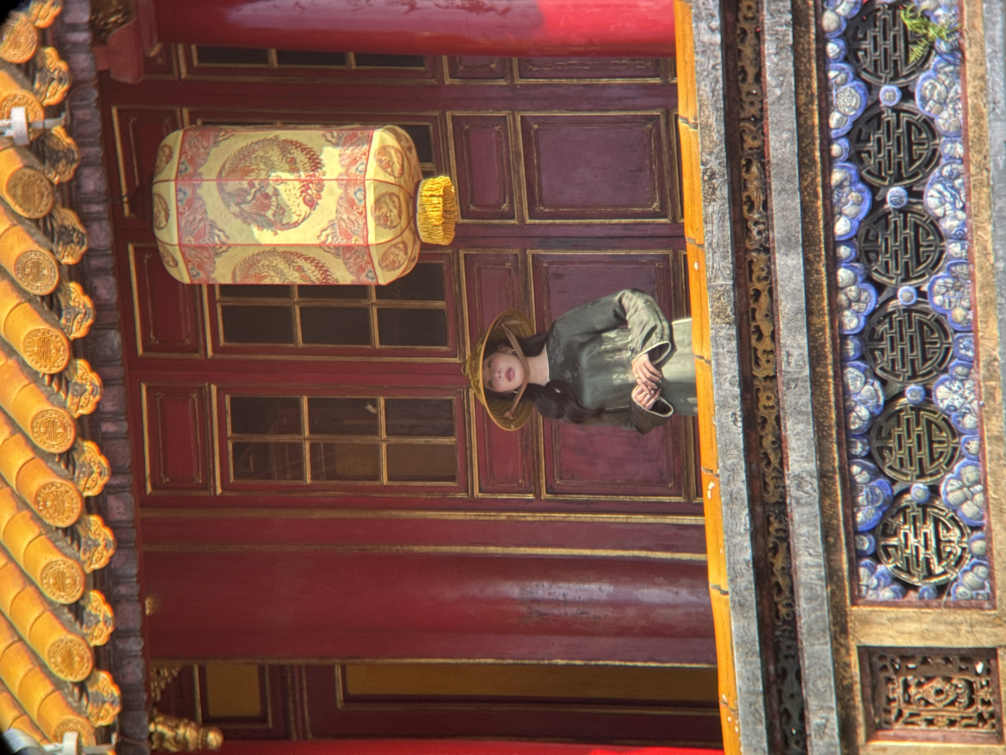 A woman wearing a conical hat and a dark green silk dress stands on a balcony in front of red doors. She is holding her hands clasped in front of her. A large yellow lantern with red phoenix designs hangs above and slightly to her right. The background is the exterior of a building with a red facade, ornate yellow roof tiles, and intricately carved decorative elements on the lower part of the frame. The scene appears to be set in the Imperial City of Huế, located in Phú Xuân Ward, Vietnam.