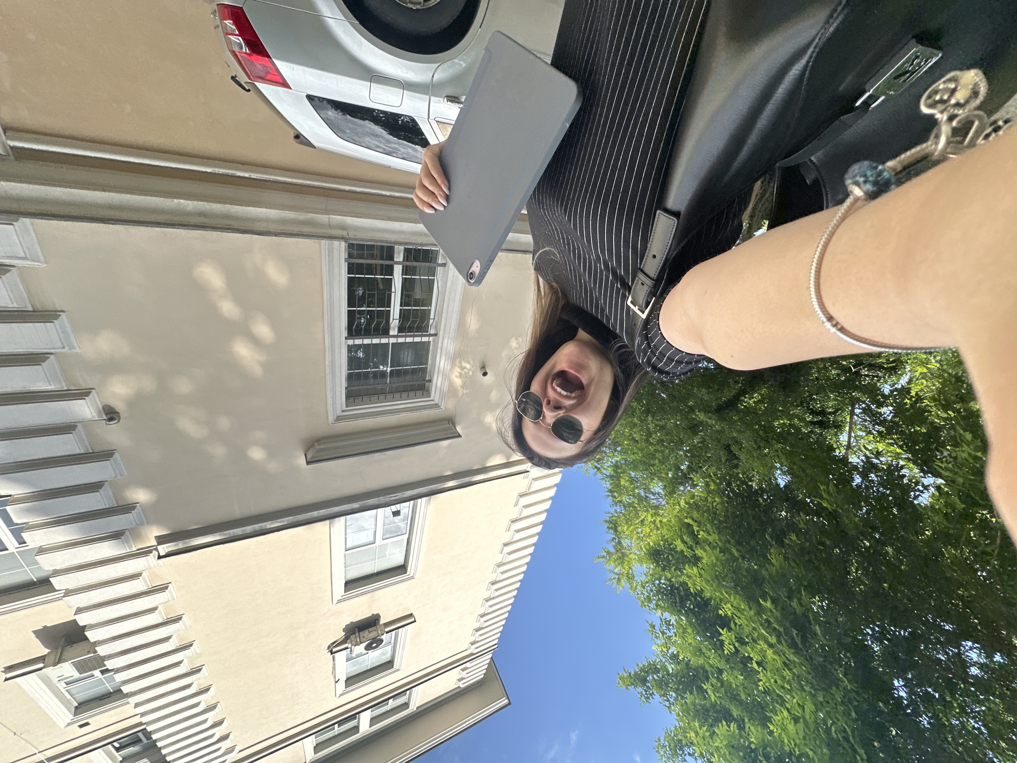 A selfie-style, rotated, full-shot image depicts a fair-skinned young woman wearing round, dark sunglasses and a black pinstripe top, holding a gray tablet and a black handbag with a silver bracelet on her wrist. Her mouth is wide open in an exaggerated expression. The background features a light beige building with architectural details such as tiered eaves and windows, a portion of a white car, trees with green leaves, and a blue sky. The location appears to be an urban setting, potentially within the city of Sofia, Bulgaria, given the architectural style.