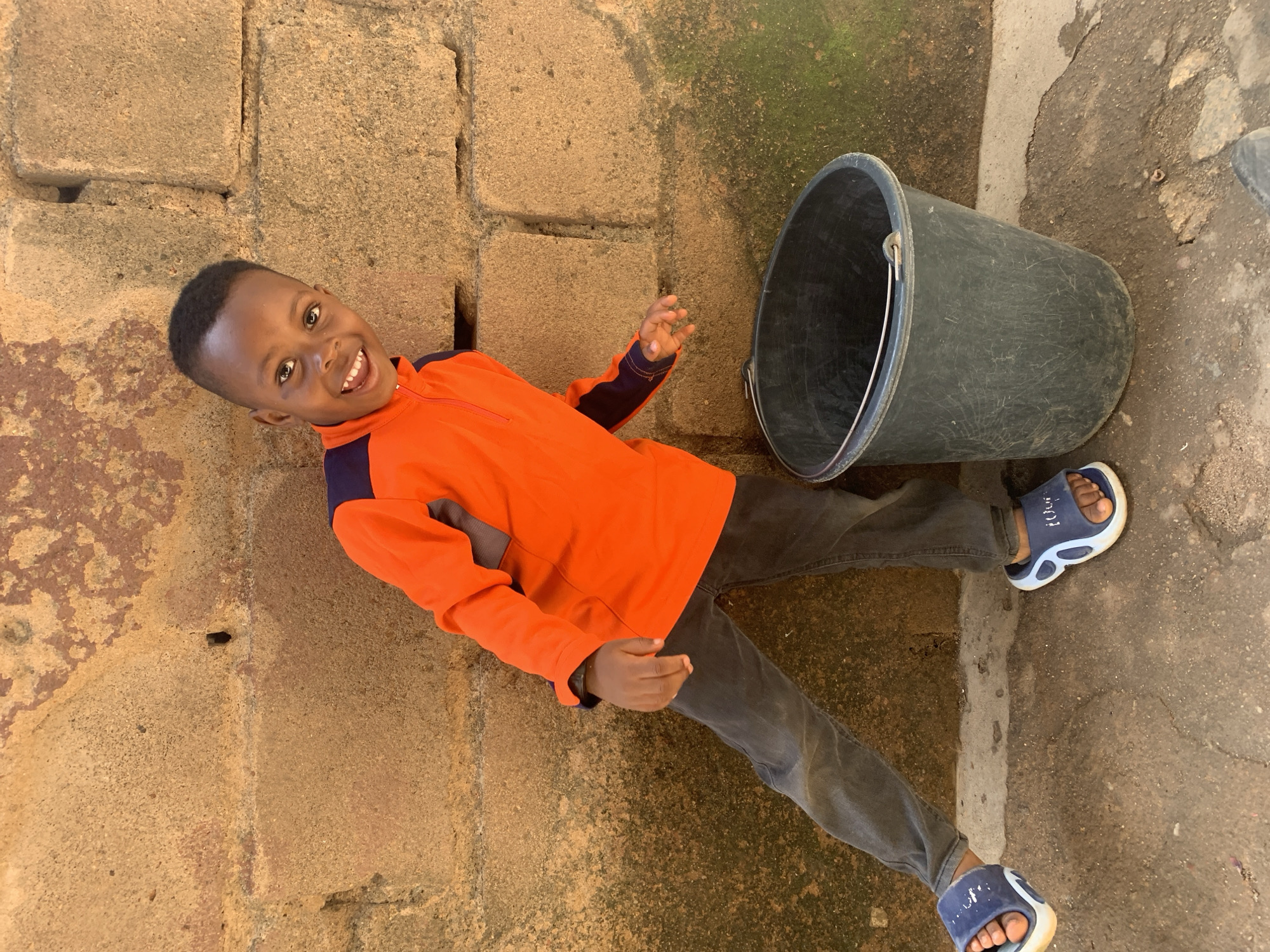 A young boy of African descent, with dark skin and short black hair, stands leaning against a rough, textured wall made of large, uneven concrete blocks. The wall is a reddish-brown color, with patches of darker discoloration and green moss in some areas. The boy is wearing an orange long-sleeved shirt with navy blue accents at the collar and shoulders, and dark gray denim pants. He has on blue and white slide sandals. To his right, a large, dark gray plastic bucket with a metal handle sits on the ground. The ground is a mix of dirt and concrete, with some small stones visible. The boy is looking towards the camera with a wide, joyful smile and his mouth open. He is positioned to the left of the bucket, with his left leg extended and his body slightly turned. The scene appears to be outdoors, in a setting that could be common in many neighborhoods in Jos, Nigeria, characterized by its simple construction materials and dry, earthy ground.