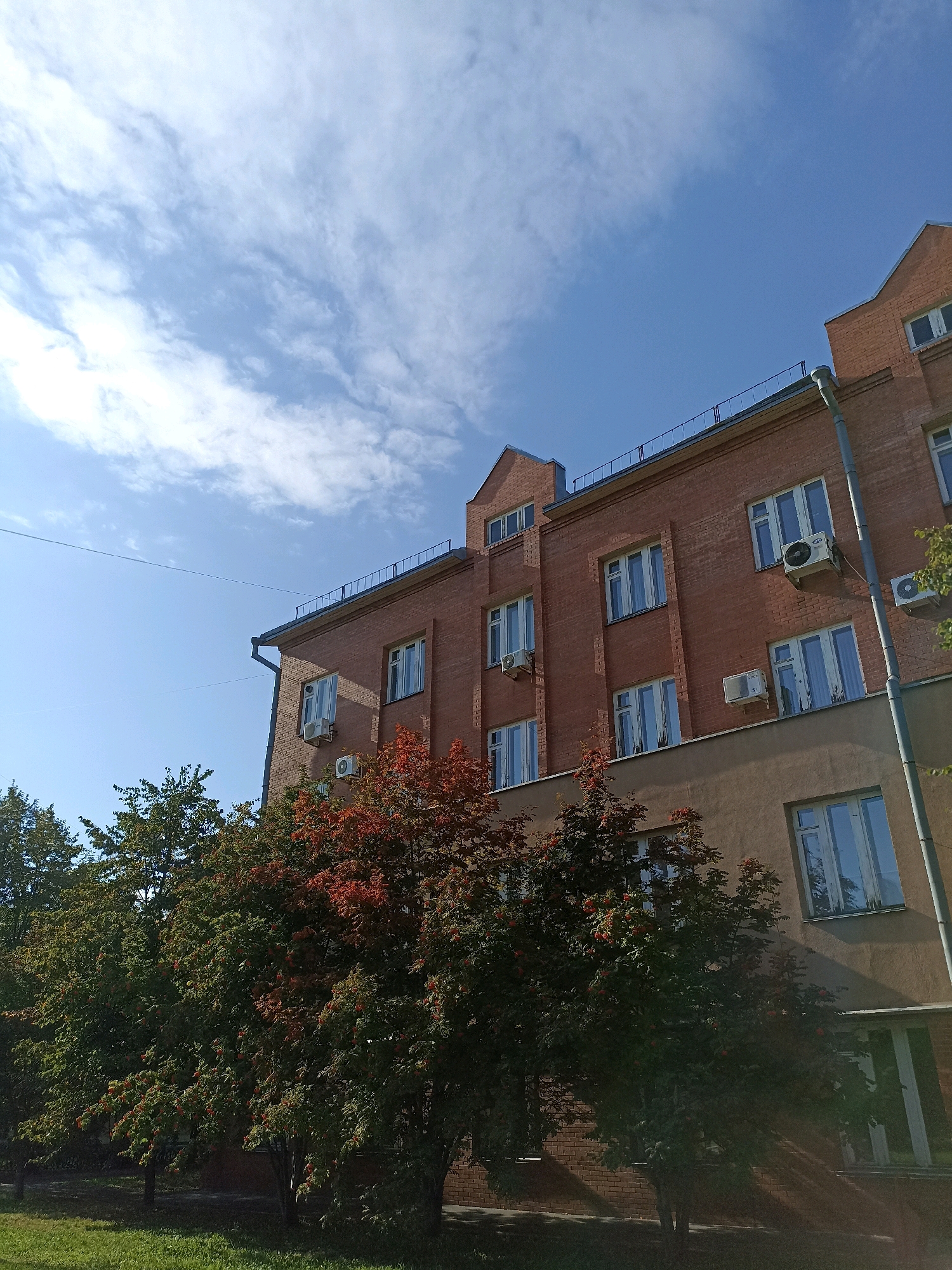 The image shows a building constructed with red brick on a sunny day with a blue sky and scattered clouds. The building has multiple windows, some with air conditioning units installed outside. Trees with green and red foliage are visible in front of the building, suggesting it might be autumn. There is a patch of green grass in the foreground. The scene appears to be located in an urban or suburban area.