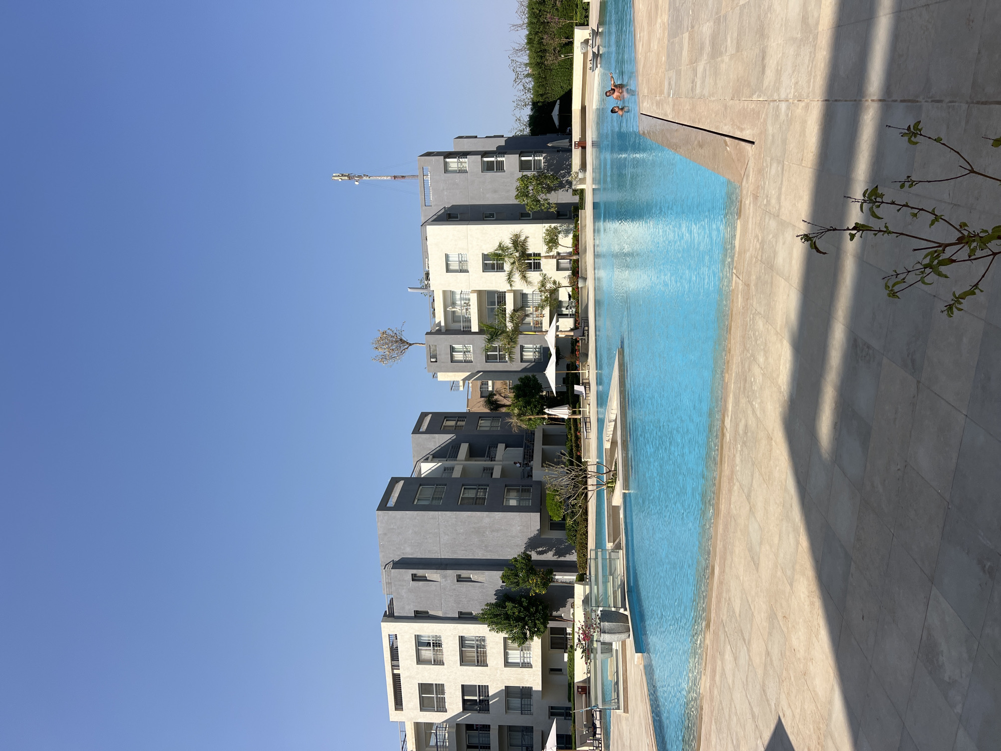 An outdoor swimming pool area is depicted on a clear, sunny day. The pool, filled with bright blue water, is a prominent feature, with a tiled deck surrounding it. In the background, modern apartment buildings with a mix of white and grey facades are visible. Several palm trees and other greenery are planted around the buildings and pool area, adding to the resort-like atmosphere. Two people are in the swimming pool, appearing to be engaged in conversation or activity. The overall scene suggests a recreational or residential complex in a warm climate.
