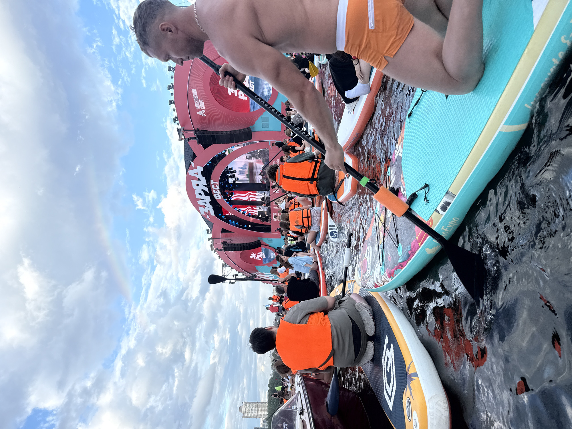 An overhead, medium shot captures a group of people on stand-up paddleboards in the water, facing a large, red stage structure adorned with lights and banners. A man in orange shorts and a necklace, holding a paddle, is in the foreground on the right, facing left. The stage features Cyrillic text, "ЗАРЯ" and "МОСКОВСКИЙ СПОРТ." A faint rainbow is visible in the cloudy sky above. The water is dark and rippling, with reflections of the stage and people. Several individuals on the paddleboards are wearing orange life vests. In the background, buildings and trees can be seen on the shoreline.