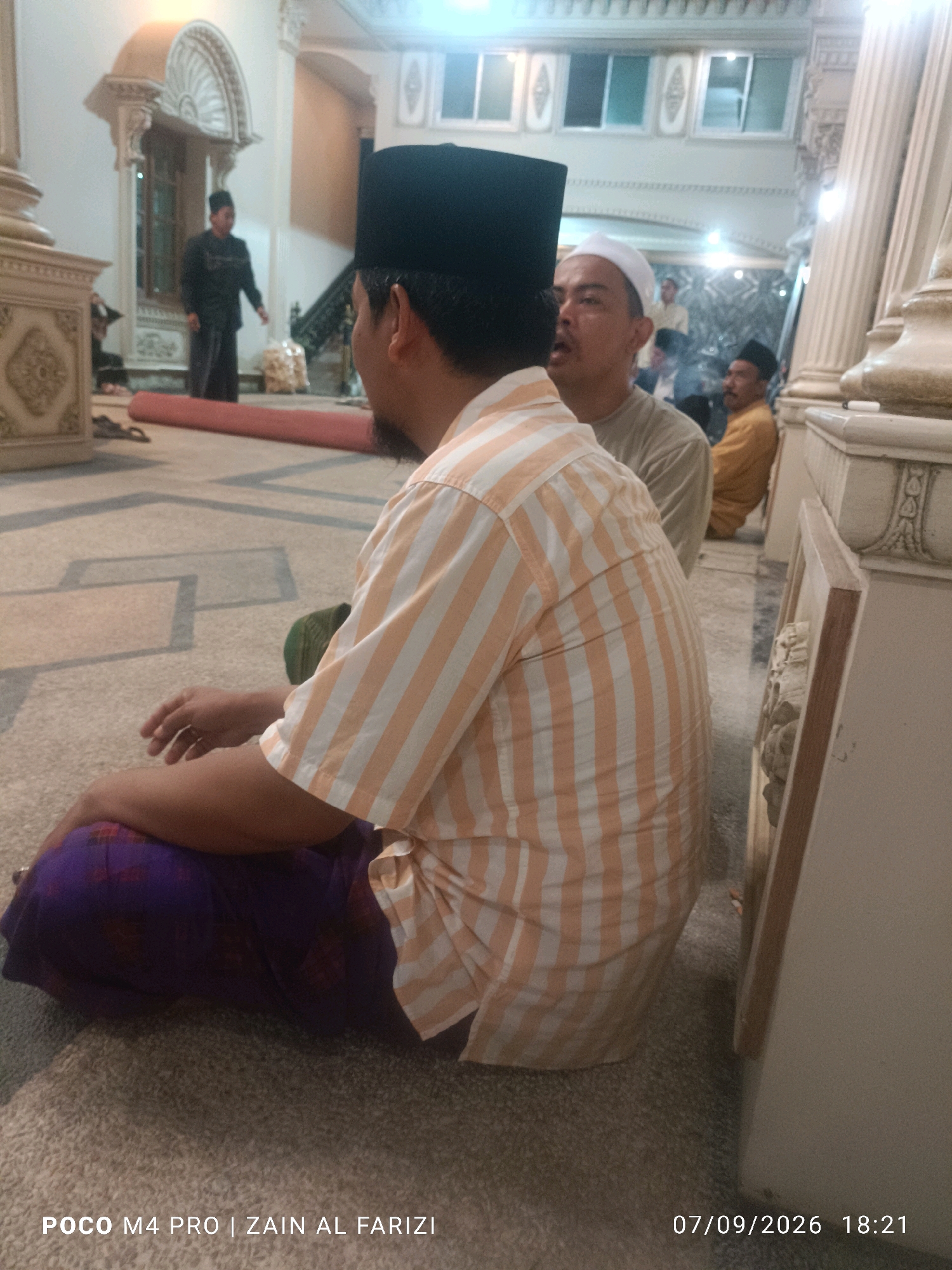 In the city of Buduran, Indonesia, an indoor scene captures individuals in what appears to be a religious or cultural gathering. The primary focus is on a man seated on the floor, facing away from the camera. He wears a black skullcap and a long-sleeved shirt with vertical stripes in orange and white. He is also wearing a dark purple sarong. To his right, another man is visible, wearing a white cap and a light-colored shirt, looking towards the seated man. Further in the background, another man in a yellow garment is seated. The environment features ornate architectural elements, including columns and arched structures, suggesting a mosque or a similar place of worship. Red rolled mats are present on the floor, possibly for prayer. The lighting suggests an interior space, possibly during the day with artificial lighting. The timestamp at the bottom right indicates the photo was taken on 07/09/2026 at 18:21.
