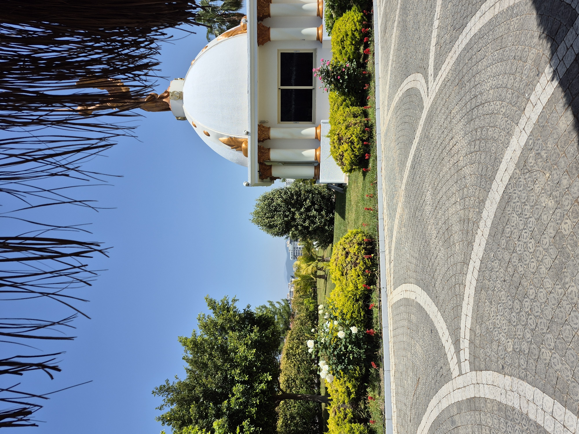A vertical outdoor shot shows a white gazebo-like structure with a rounded roof and gold decorative accents in Alanya, Turkey. The structure is bordered by green shrubs with red and pink flowers. The ground is covered in gray pavers with white arc patterns. Blue sky is visible in the background, framed by the thatched roof in the foreground. Lush greenery and trees are visible in the distance. Buildings are also visible in the distant horizon.
