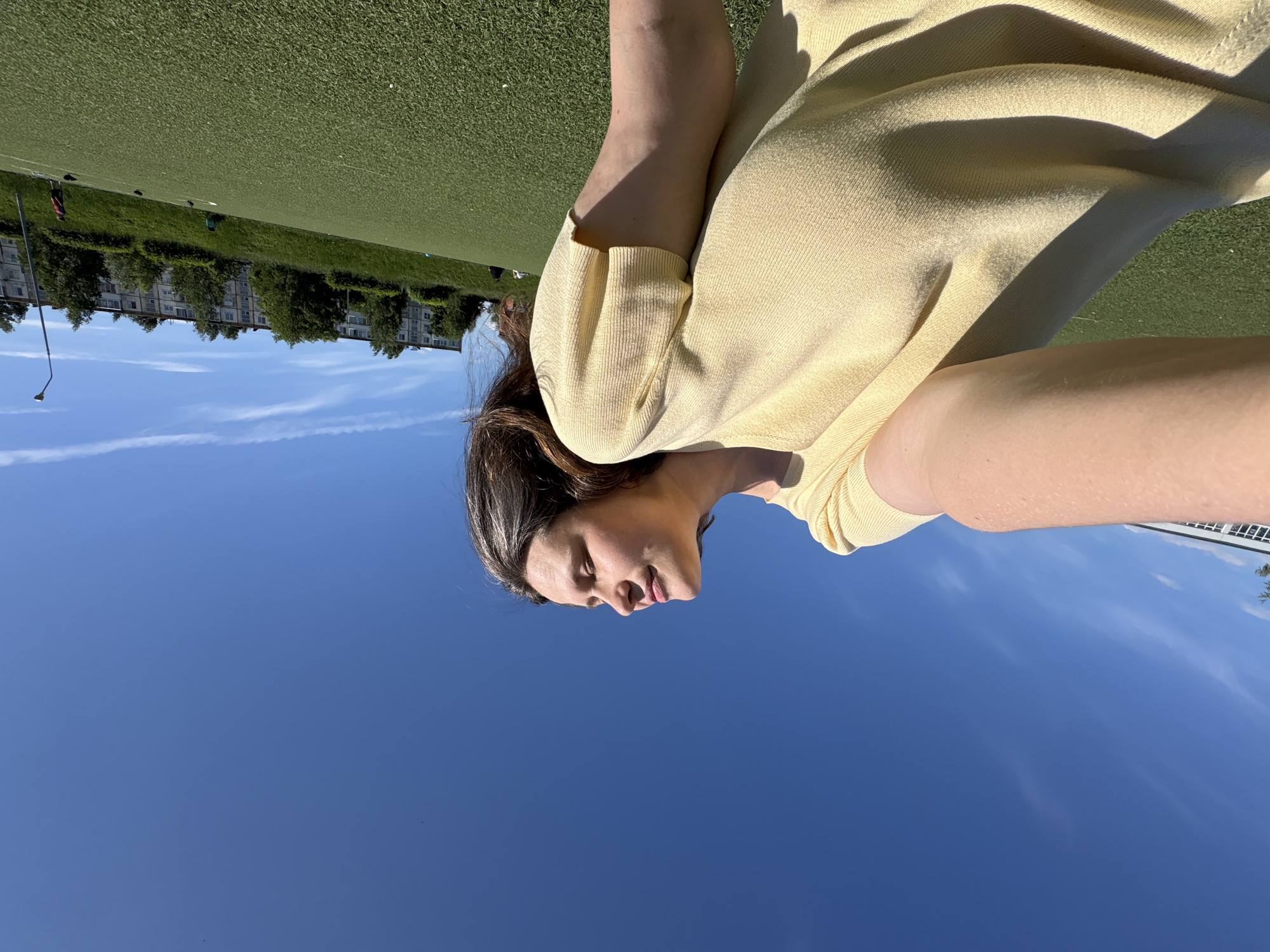 This low-angle photograph depicts an adult female positioned on an artificial green turf field, under a clear blue sky, in an urban environment identified as Chaikovsky, Russia. The woman, occupying the left side of the frame, is wearing a light yellow, short-sleeved knit top. Her head is tilted significantly upwards, with her eyes appearing closed or partially closed, suggesting direct exposure to sunlight. Her dark hair is visible falling over her shoulder on the right. Her left arm is extended and visible in the lower left quadrant of the image.
The foreground and midground consist of artificial green turf, indicative of a sports field, with faint white lines discernible on its surface. Several dark-colored birds, consistent with pigeons, are present on the turf in the mid-distance. The background features a gently sloping grassy hill covered with numerous green trees and shrubs. Above this natural incline, a long, multi-story residential building, characterized by its uniform architectural style and multiple windows typical of Soviet-era construction, spans across the horizon. A street lamp post stands vertically on the right side of the frame, extending above the building line. Scattered individuals are visible in the distant background on the field, one wearing a red shirt. A blue waste receptacle is located on the grassy slope of the hill.
The sky is bright blue and largely cloudless, with a few sparse, wispy white clouds present in the upper portions of the frame. The scene is illuminated by strong direct sunlight, casting distinct shadows on the woman's face and clothing, as well as on elements within the environment.