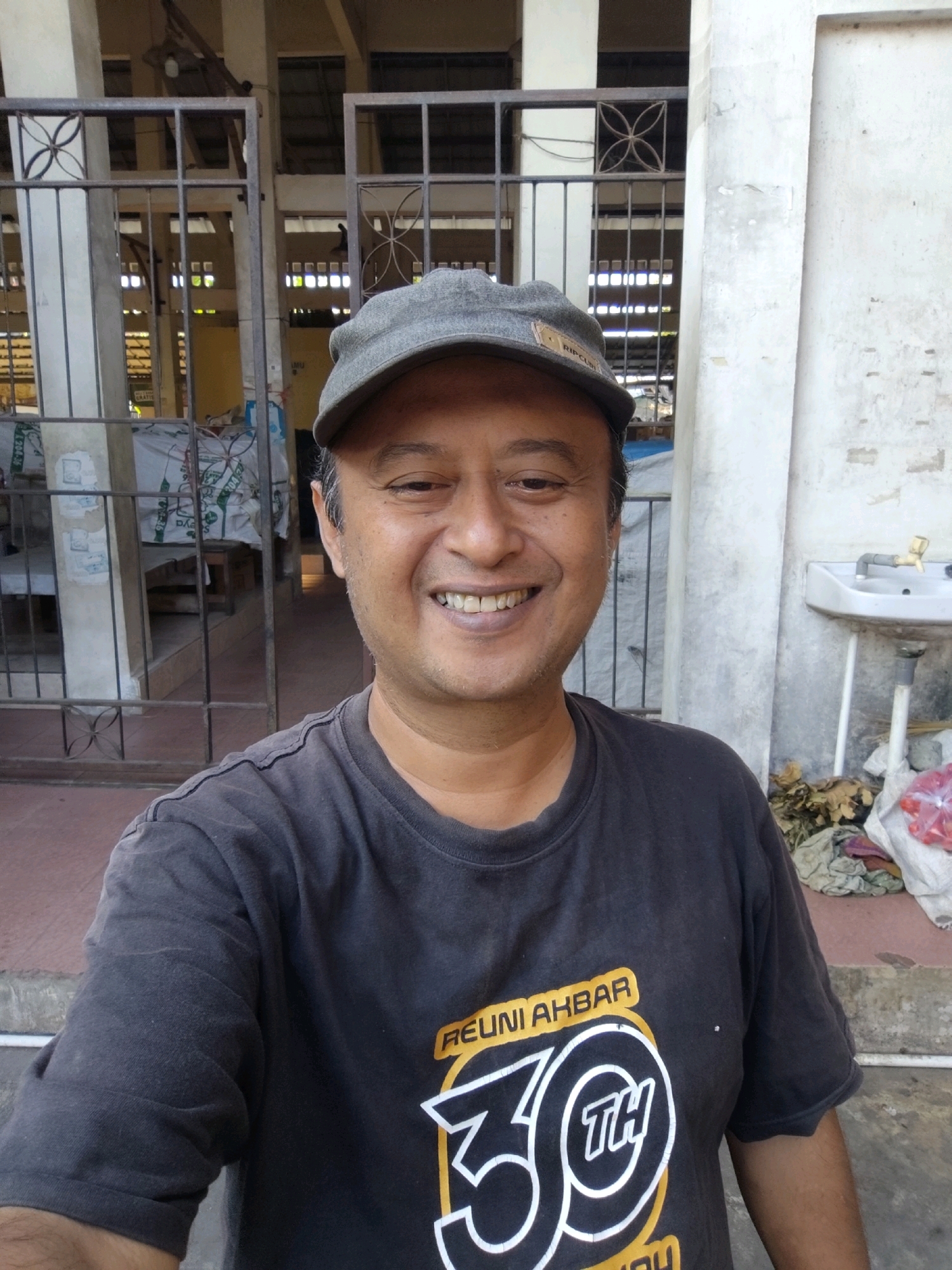 A middle-aged East Asian man, wearing a grey baseball cap and a dark grey t-shirt with a graphic that reads "REUNI AKBAR 30TH", smiles at the camera. He is standing in front of a building with a white wall and some metal gates. Behind him, there are some bags of goods and a sink with a faucet. The environment appears to be an outdoor market or a public space in Sanden, Indonesia. The man's facial features are visible, including his smiling mouth and eyes. The lighting is bright, suggesting it is daytime.