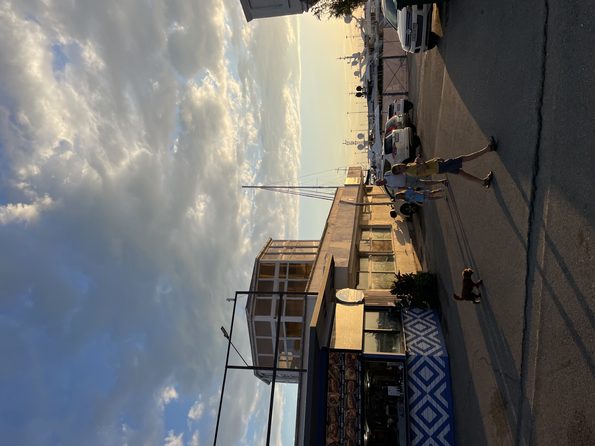 The image depicts a scene in Sochi, Russia, at either sunrise or sunset, as indicated by the warm, golden light and long shadows cast across the asphalt. The sky is filled with dramatic, textured clouds, transitioning from a bright, illuminated yellow at the horizon to a deeper blue and gray overhead.
In the foreground, a family of three, likely two adults and a child, are walking away from the camera towards the right side of the frame. They are dressed in casual summer clothing. Behind them, a dog, appearing to be a golden retriever or similar breed, trots along the pavement.
The setting appears to be a waterfront promenade or a street adjacent to a marina. Several white yachts are docked in the background, their masts and rigging visible against the sky. A building with large glass windows and a textured facade runs along the left side of the image, housing a business with a menu displayed on a digital screen, showing various food items. A striking blue and white geometric pattern adorns the lower part of the building. The overall mood is serene and summery, suggesting a leisurely stroll in a coastal town.