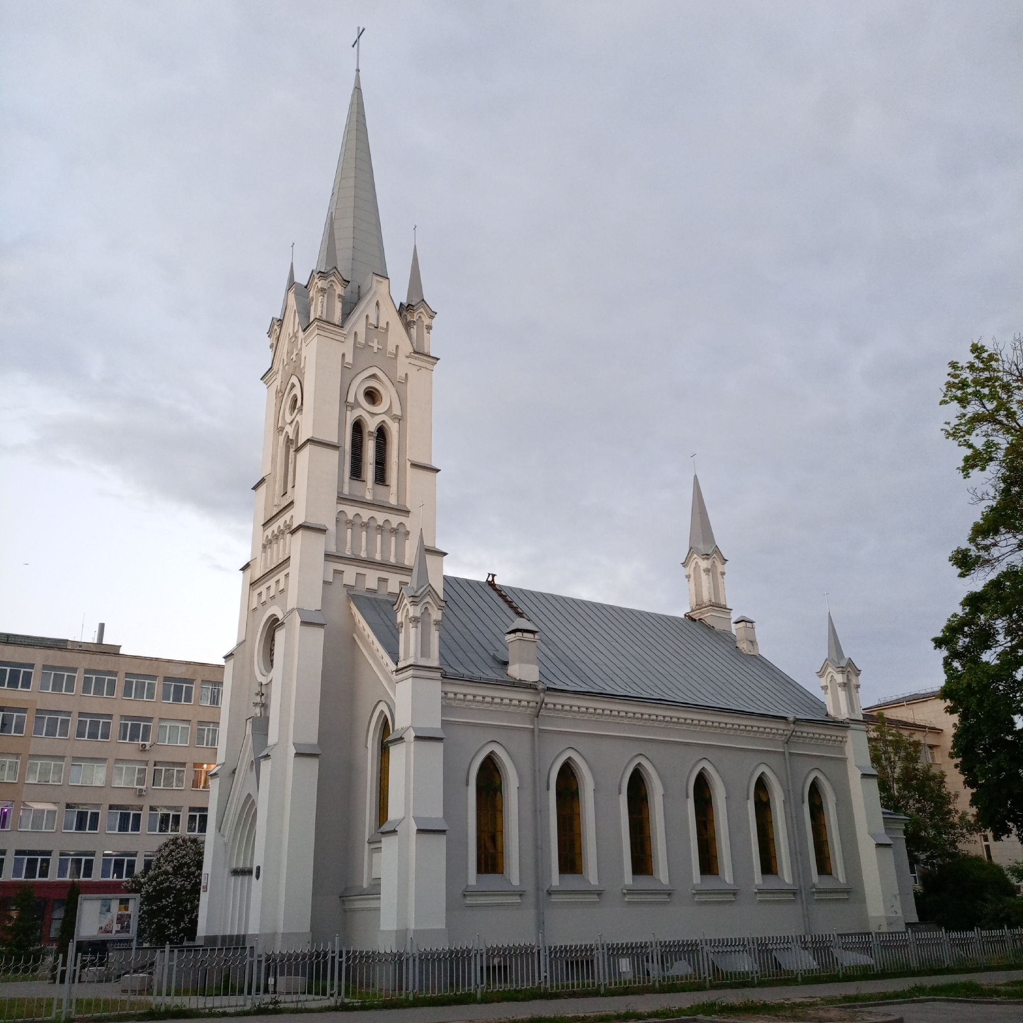 A low-angle, medium shot shows a gray church with a tall, white spire in the city of Unknown, Unknown. The church has a gray roof and many arched windows. A tall, white spire rises from the left side of the church. The top of the spire is capped with a cross. A gray fence surrounds the church. A large building with many windows is visible behind the left side of the church. Trees are visible behind the right side of the church. The sky is overcast.