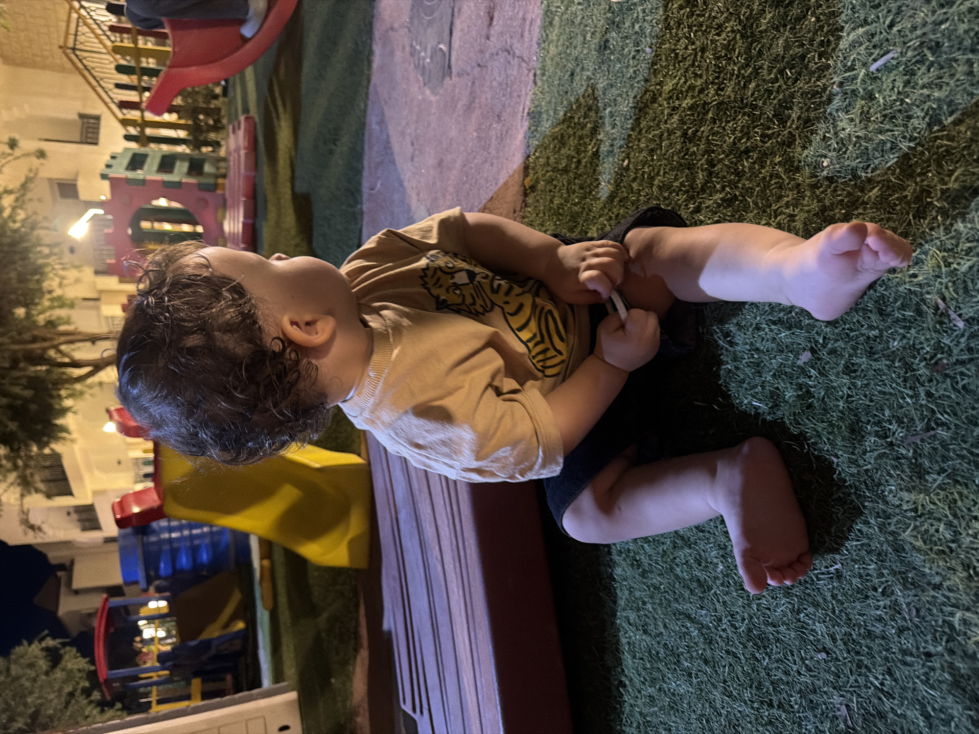 A toddler sits on artificial grass in a playground at night. The toddler, facing away from the camera and to the right, wears a tan t-shirt with a tiger graphic and dark shorts. The toddler's left foot is visible, resting on the grass. The background shows a colorful playground with slides, climbing structures, and buildings illuminated by artificial lights. A person can be seen in the far right background. The location is specified as Sharm El Sheikh, Egypt.