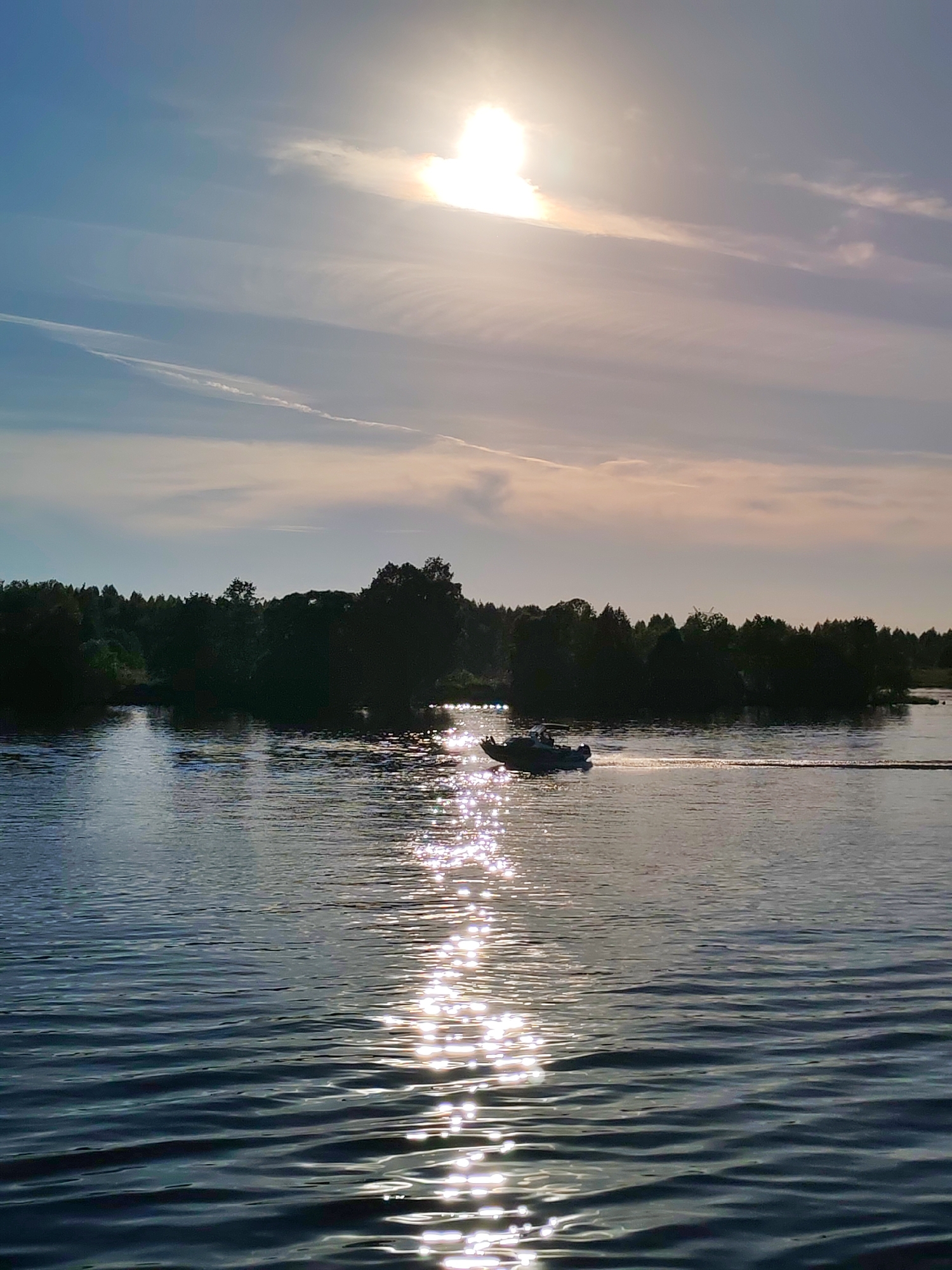 The image depicts a tranquil aquatic landscape under a bright, late afternoon or early evening sky, specifically located in Golovino, Russia. A prominent sun, high in the upper-right quadrant, casts a brilliant glare directly across the water's surface, creating a sparkling, elongated reflection extending towards the foreground. The sky is a gradient of light blue to pale orange near the horizon, populated by wispy, horizontally stretched clouds and a faint, linear contrail in the upper-left.
A dense, dark green treeline forms the distant bank, silhouetted against the lighter sky and water. On the water, a single dark-hulled motorboat, possibly an inflatable type with an outboard motor, traverses from the left towards the right of the frame, leaving a distinct wake behind it. The boat is positioned within the sun's reflected path on the water, with its details largely obscured by shadow and distance. The water itself exhibits subtle ripples, particularly in the foreground and areas not directly illuminated by the sun's reflection. No individuals are clearly discernible on the boat or shore.